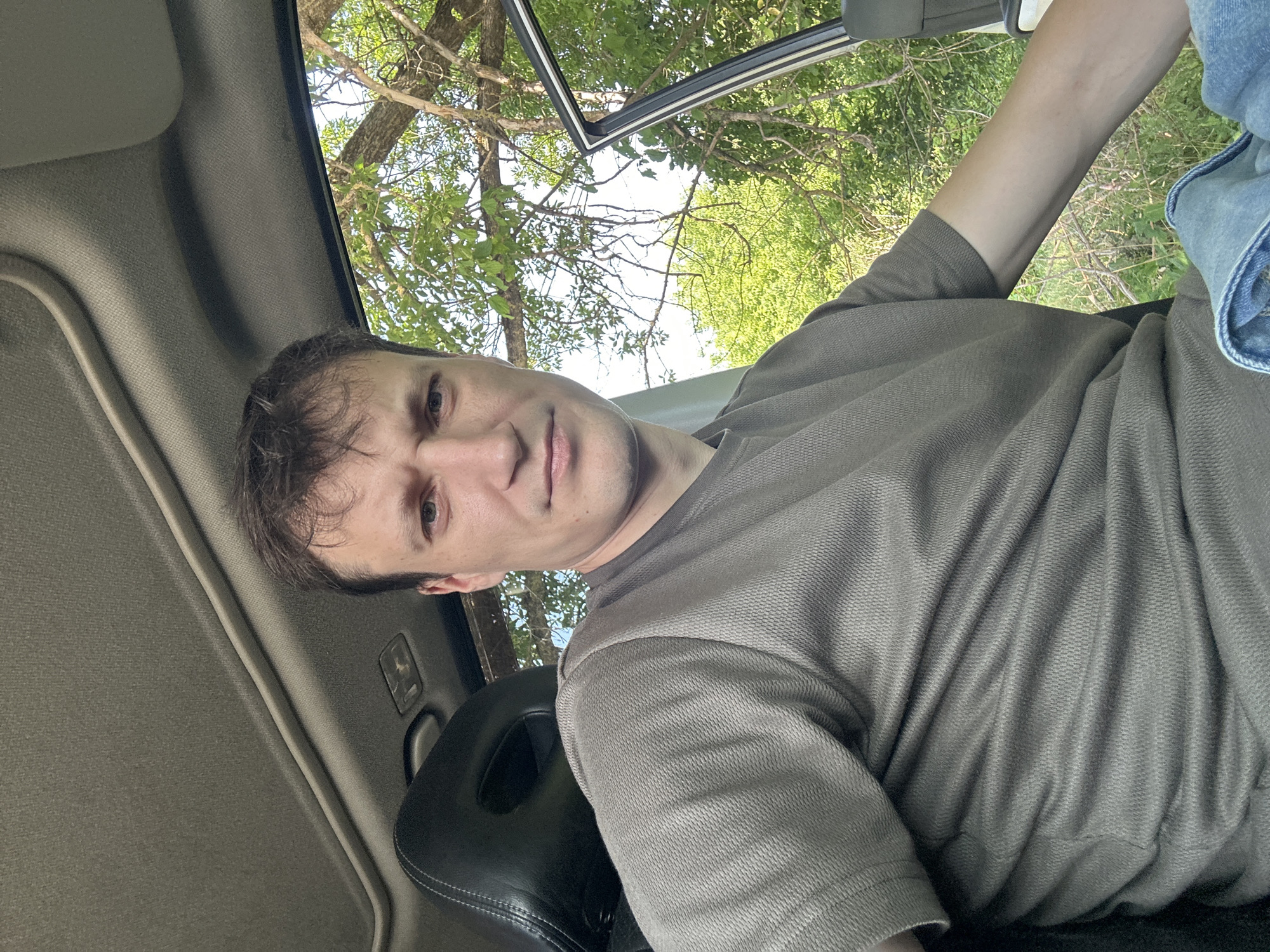 A man is sitting inside a car, with the driver's side door open. He is wearing a grey t-shirt and blue jeans. The car is parked outdoors, with trees and foliage visible in the background. The image was taken during the daytime, as indicated by the bright sunlight. The location is Makiv, Ukraine.