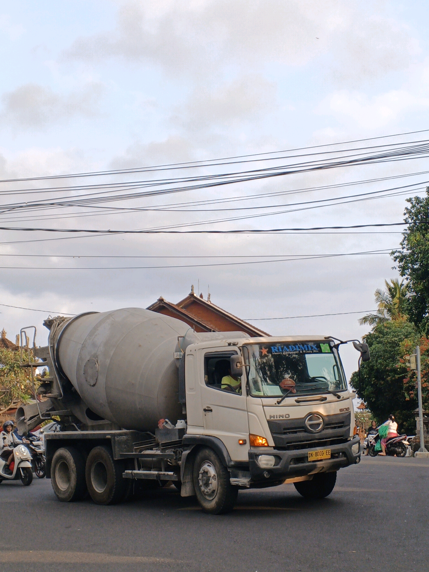 In Sukawati, Indonesia, a white Hino cement mixer truck is driving on a paved road under an overcast sky with visible power lines overhead. The truck has a large, gray cement drum on its back. The driver, wearing a yellow shirt, is visible in the cab. To the left of the truck, a person on a white scooter is riding past. In the background, there are buildings with traditional Balinese architecture, lush green trees, and more power lines. To the right of the truck, another person on a scooter, wearing a green garment, is also visible. The truck's license plate reads "DK 8000 EE".
