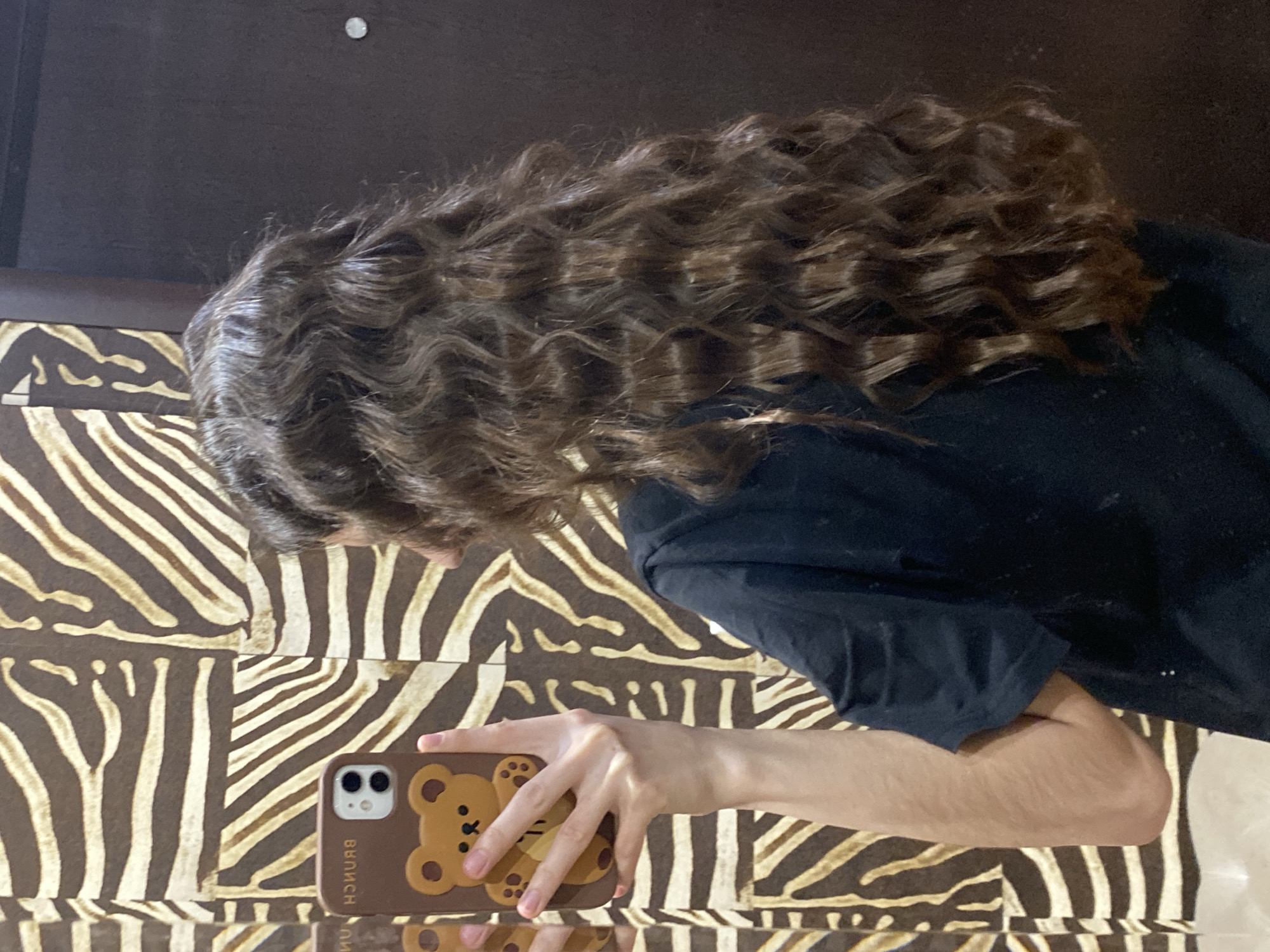 A person is seen from an overhead, slightly angled perspective. The person has long, brown, curly hair that cascades over a patterned surface. The person is wearing a dark-colored shirt. The person is holding a smartphone with a brown case adorned with a bear design. The patterned surface appears to be a zebra-striped surface. The location cannot be determined from the image. The image does not provide sufficient information to locate it in Votkinsk, Russia.