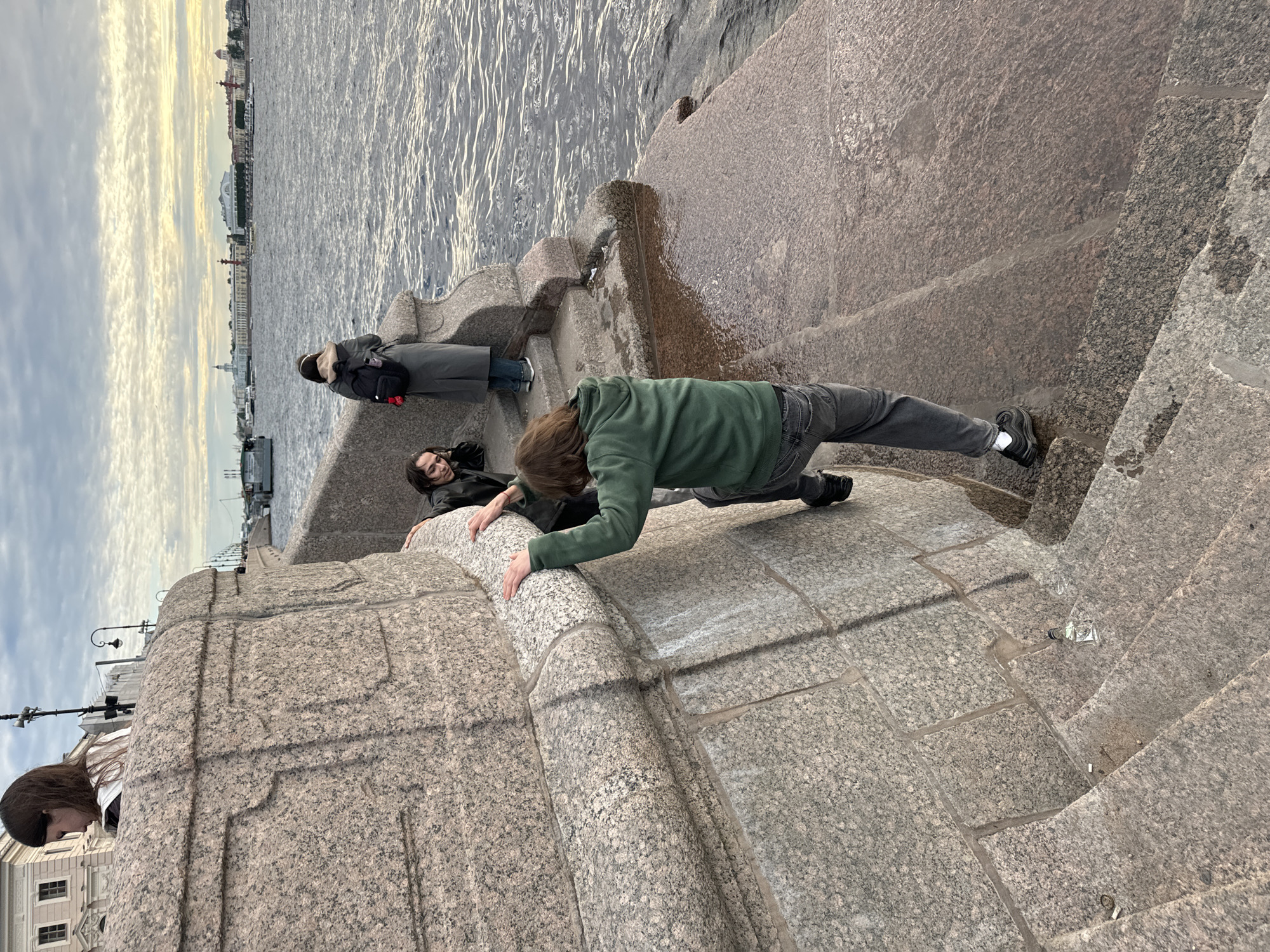 The image depicts a segment of the embankment of the Neva River in Saint Petersburg, Russia, likely near the Hermitage Museum, given the architectural style. The foreground features a massive granite embankment with stairs leading down to the water. A young person with long brown hair and wearing a green hooded sweatshirt and grey jeans is in the process of climbing up the embankment, gripping the stone with their hands and feet. Another individual with dark hair and a black jacket is partially visible on the stairs, looking towards the person climbing. Further in the background, a person in a long grey coat with a backpack is standing by the river, looking out at the water. The river is choppy with moderate waves, and the sky is overcast with hints of yellow and orange light from a setting sun. The embankment itself is wet in places, with puddles of water on the stairs and a sheen on the granite. In the distance, the skyline of Saint Petersburg is visible, with classical buildings and bridges. The overall atmosphere suggests a cool, possibly autumnal evening.