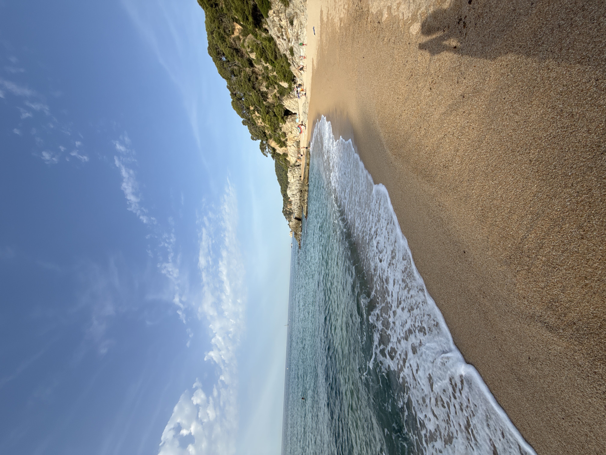 This is an eye-level, long shot of a beach in Lloret de Mar, Spain. The beach is sandy and slopes gently towards the sea. The sea is a clear blue-green with small waves lapping at the shore. There are some rocks and vegetation on the left side of the image, and a hill covered in trees on the right. In the distance, there are a few people on the beach, some swimming in the sea, and a sailboat on the horizon. The sky is blue with some scattered clouds. The overall mood is peaceful and relaxing.