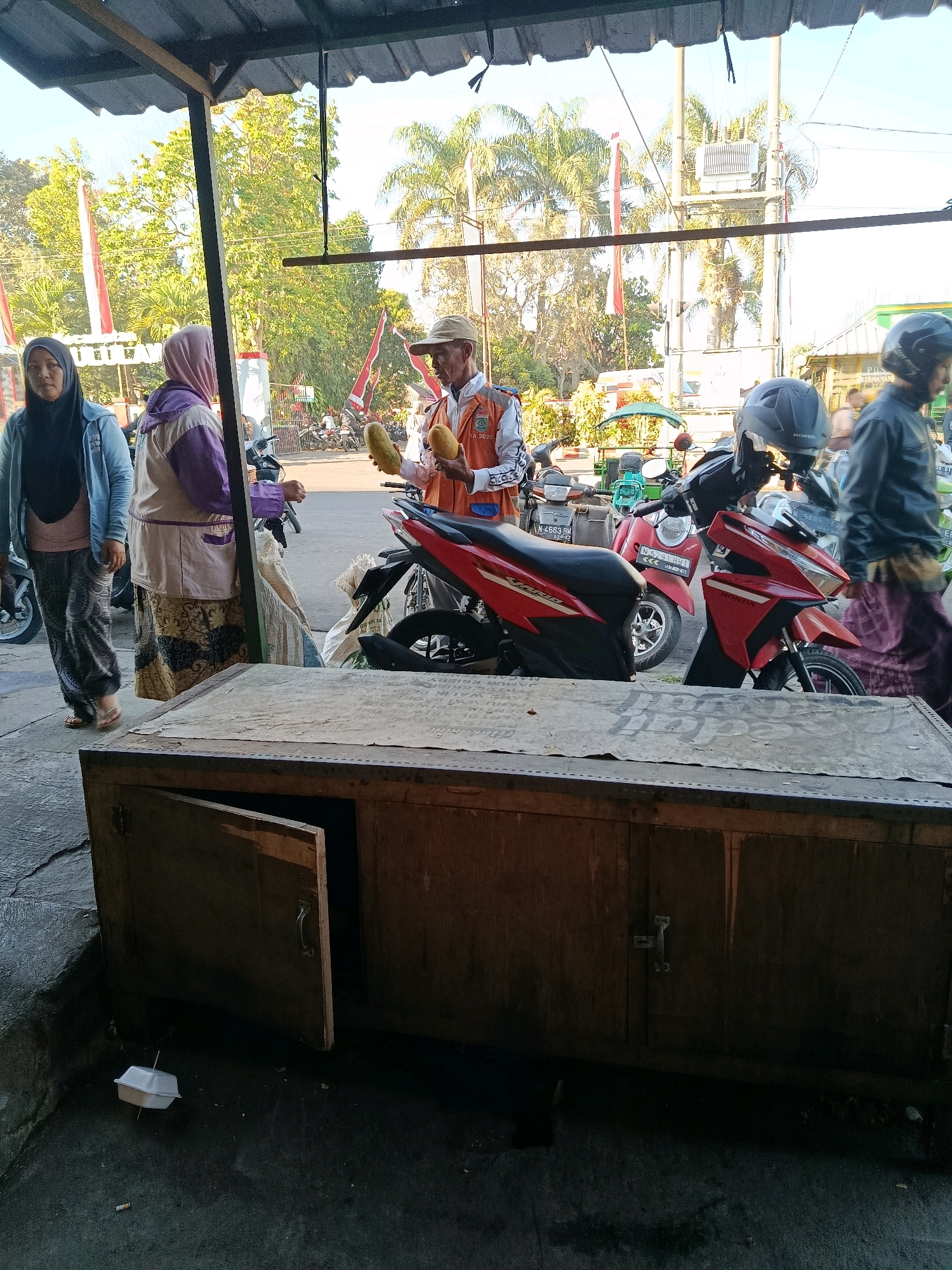 This image captures a street scene in Bululawang, Indonesia. In the foreground, a weathered wooden cabinet with one door ajar sits on a paved surface. A white styrofoam container with a stick impaled in it and a cigarette butt are on the ground near the cabinet. Behind the cabinet, several people are visible. A woman wearing a black hijab and a light blue jacket stands to the left, looking towards the viewer. Next to her is another woman in a purple and cream jacket and a patterned skirt. A man in a hat and an orange vest is in the center, holding two light brown, elongated objects that appear to be bananas or similar fruit. He is interacting with the women. Several motorcycles are parked behind the people, and further in the background, there are trees, buildings, and utility poles under a bright sky. The overall setting suggests a casual outdoor marketplace or roadside stall.
