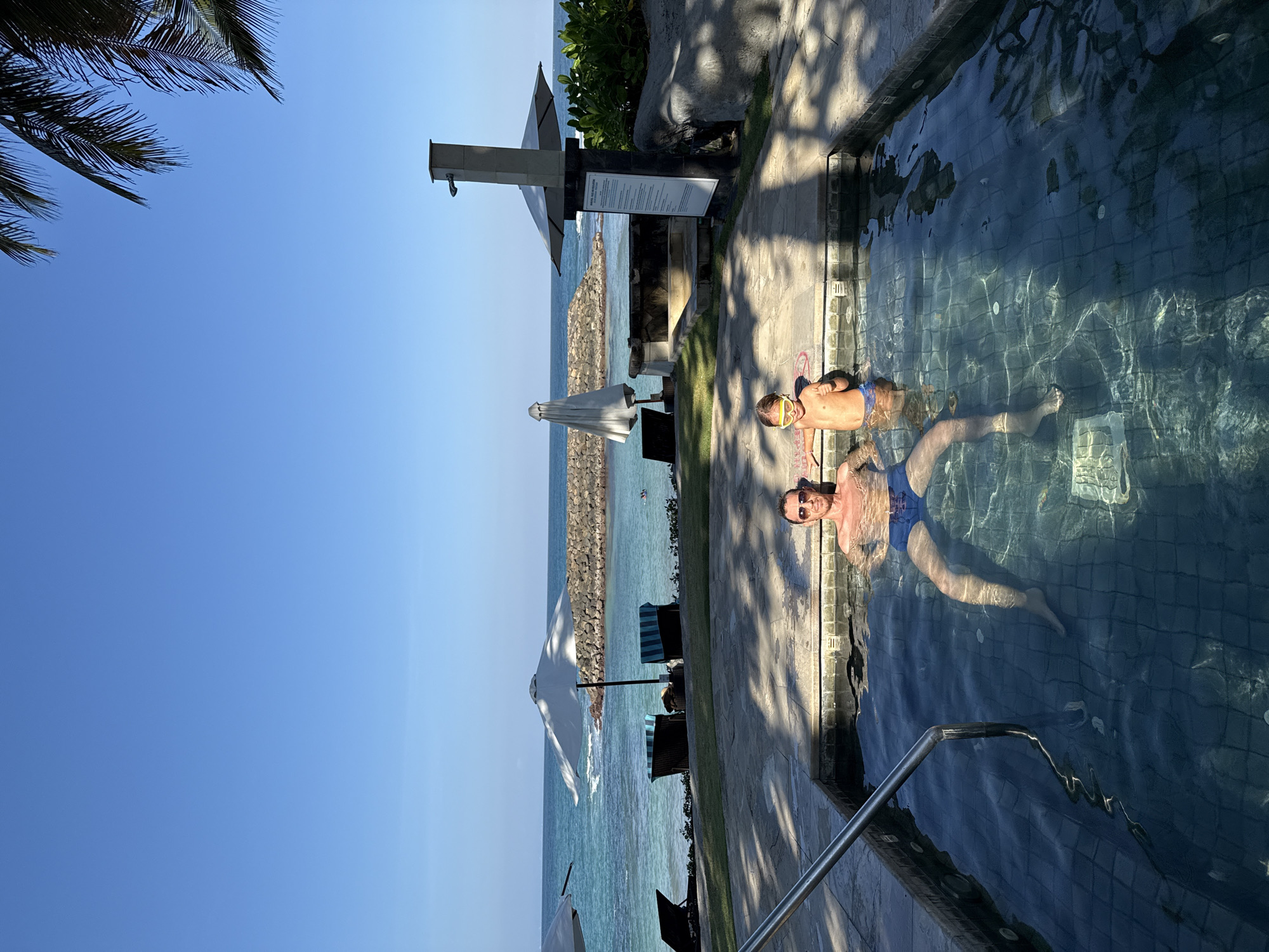 In Kuta Selatan, Indonesia, a man and a young child relax in a tiled hot tub with clear blue water. The man, wearing blue swim trunks, is reclined with sunglasses on, while the child, in blue swim trunks and yellow goggles, gives a thumbs-up. The hot tub is set on a stone patio surrounded by lush greenery and palm trees. In the background, a tranquil turquoise ocean meets a clear blue sky, with a man-made rock barrier extending into the water. Beach chairs and umbrellas are visible along the shoreline, suggesting a resort or hotel setting. The sun casts dappled shadows across the patio, indicating it is daytime.