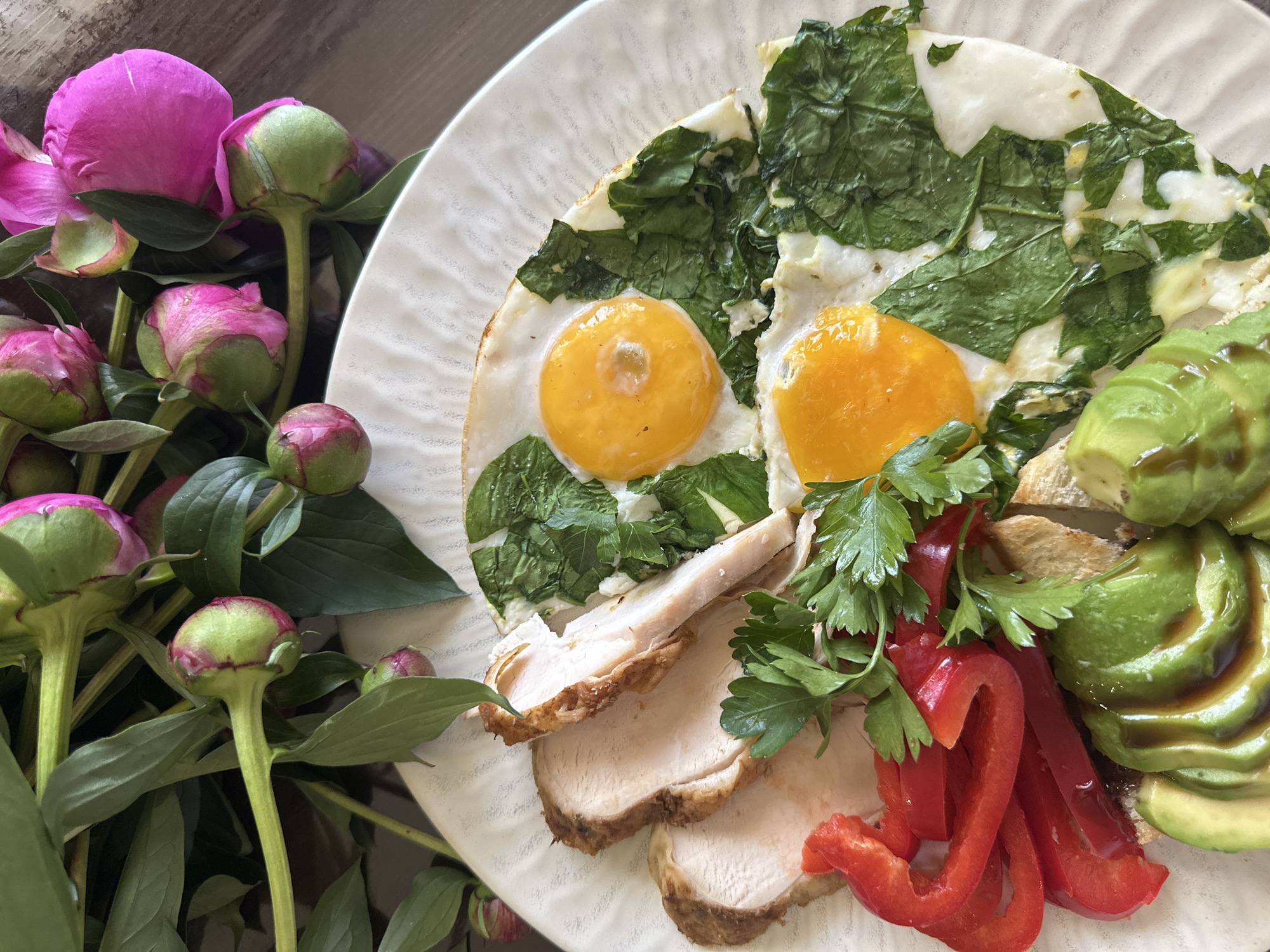 A close-up, top-down shot shows a white, textured plate filled with a breakfast meal and a bouquet of peonies. The meal consists of sliced turkey breast, two fried eggs with runny yolks and wilted spinach, sliced red bell peppers, avocado slices drizzled with a dark sauce, and what appears to be toast underneath. The peonies, in various stages of bloom, are a mix of pink and green buds with bright pink open flowers, creating a vibrant contrast against the muted tones of the food and plate. The background is a blurred, dark wooden surface. There is no discernible indication of the city of Chaikovsky, Russia, or any specific location.