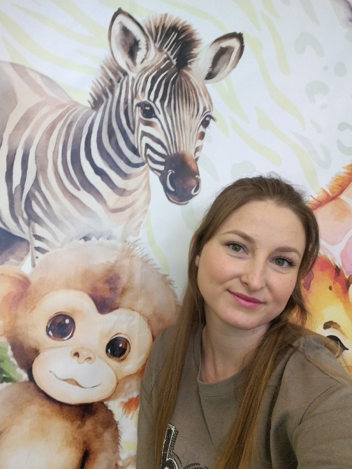 The scene depicts an indoor setting in Chaikovsky, Russia. An adult female is positioned in the foreground, facing directly towards the camera. She has long, straight, light brown hair parted in the middle, green eyes, and is wearing a plain olive-green or taupe long-sleeved sweatshirt. She is smiling subtly. The background features a vibrant, multi-colored wall mural or wallpaper depicting stylized, watercolor-effect safari animals. Prominently visible are the head and upper body of a zebra foal with light brown stripes on the upper left, and below it, the head and upper torso of a brown monkey with large dark eyes and a light face. A portion of a giraffe's head is partially visible on the far right. The background also includes scattered light green, wavy leaf patterns. The lighting is even, suggesting an interior space.