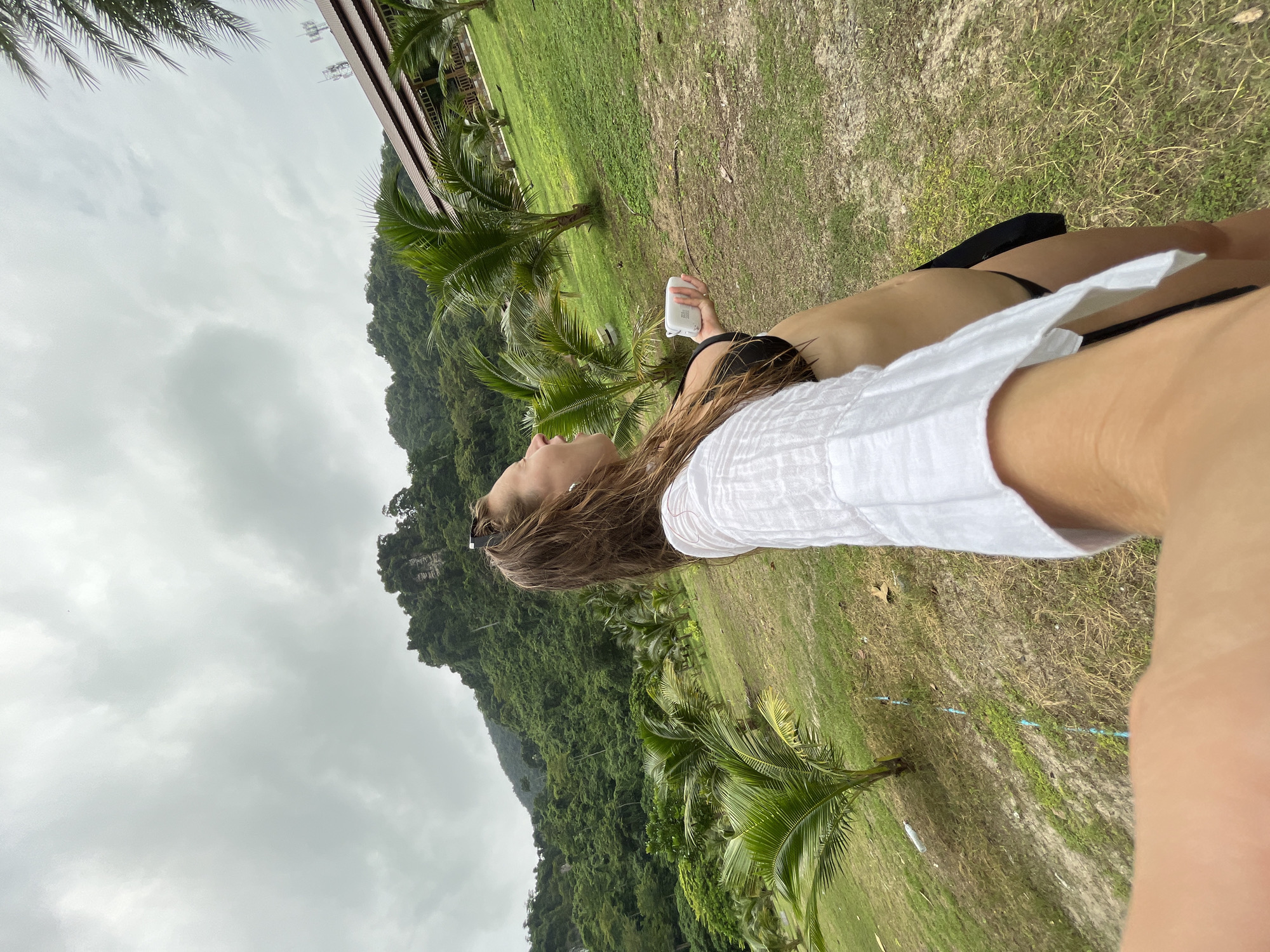 This media file is a photograph taken outdoors in Ao Nang, Thailand. The image features a person from the chest up, lying down on a grassy area. The person is wearing a white shirt and black bikini bottoms. Their head is tilted back, and they are holding a white object, possibly a sunscreen bottle, in their right hand. The background consists of a lush, green hillside covered in trees, likely part of the natural landscape of Ao Nang. In the foreground, palm trees are visible, adding to the tropical setting. The sky is overcast, suggesting cloudy weather. The overall scene evokes a sense of relaxation and vacation. There is no visible text in the image.