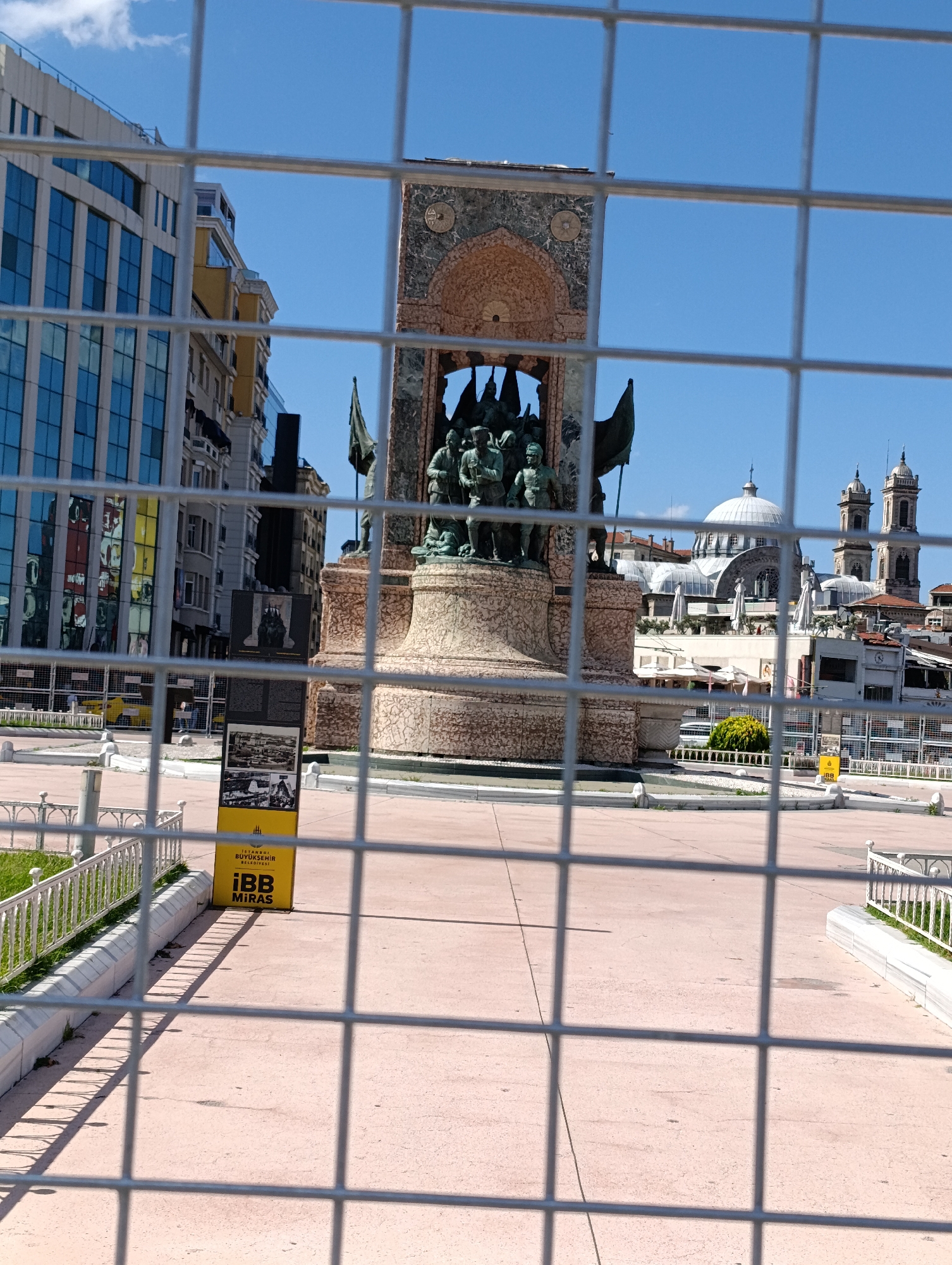 This is a medium shot, taken from behind a metal grate, of Taksim Square in Beyoğlu, Istanbul, Turkey. The monument in the center of the square, the Republic Monument, is prominently featured, with its bronze sculptures of figures and flags visible. To the left of the monument, modern buildings with glass facades reflect the blue sky and clouds. To the right, a white-domed building with two towers, possibly a mosque, is partially visible. The ground of the square is paved with pinkish material. A yellow informational sign with Turkish text and images is placed on the ground to the left of the monument. A white metal fence lines the edge of the square in the foreground. The overall impression is of a sunny day in a public urban space, with a sense of historical and modern architecture coexisting.