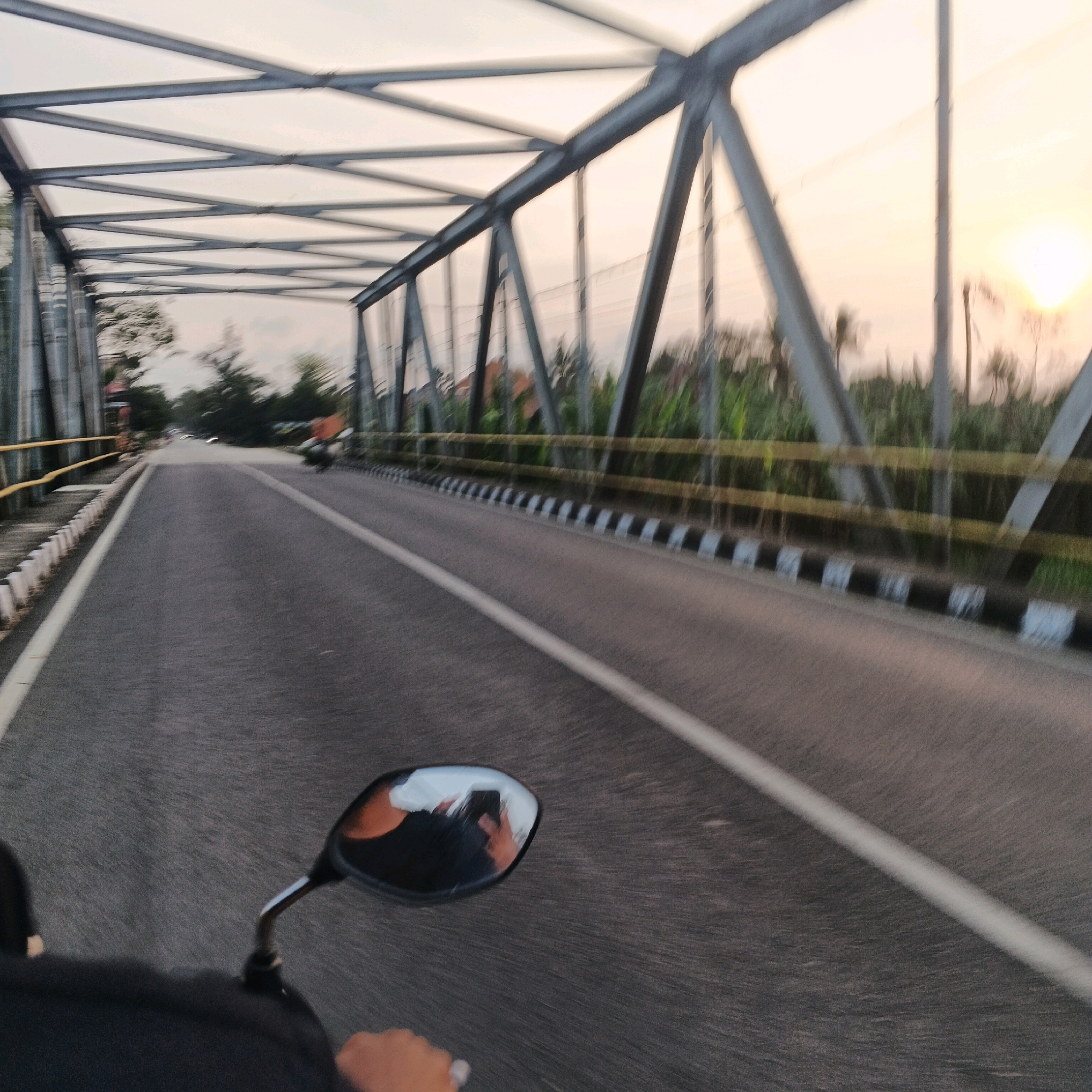 The image depicts a view from the perspective of someone riding a motorcycle on a bridge at sunset. The bridge has a metal truss structure overhead. The road is paved asphalt with a white line marking the lane. To the right of the road, there is a yellow railing and a black and white striped barrier. Vegetation, including trees and tall grass, lines the sides of the road beyond the barrier. The sky is a soft orange and yellow, indicating the sun is low on the horizon. In the distance, there are a few buildings and other vehicles on the road. A motorcycle is visible in the side mirror, reflecting the rider and the surrounding scene. The motion blur suggests the motorcycle is in motion. The location is identified as Nusajati, Indonesia.
