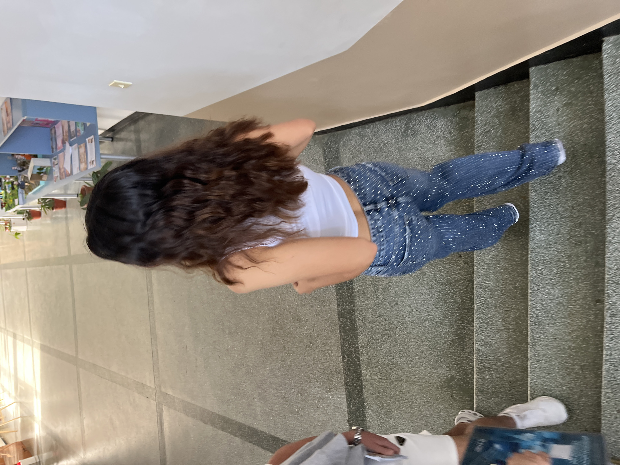The image, captured from a high angle looking downwards, shows a person standing or walking on a staircase inside what appears to be an indoor public or institutional building in Zlatograd, Bulgaria. The main subject, viewed from behind, has long, dark, curly hair and is wearing a white tank top and blue jeans adorned with numerous small white embellishments or dots. White sneakers are visible on their feet.
The staircase is constructed from a grey, speckled material, likely concrete or stone. To the left of the stairs is a light-colored tiled floor, and to the right is a smooth, beige-colored wall or railing that follows the curve of the stairs. In the background, further to the left, a blue table or shelf holds various items, including several small potted plants and what appear to be papers or display materials.
At the very bottom of the frame, partially visible, is another person. Their hands are holding what appears to be a blue folder or device. They are also wearing a white top and white sneakers, suggesting they are either on the stairs or the adjacent floor. The scene is brightly lit, indicating it is likely daytime. No specific text is clearly discernible in the image.