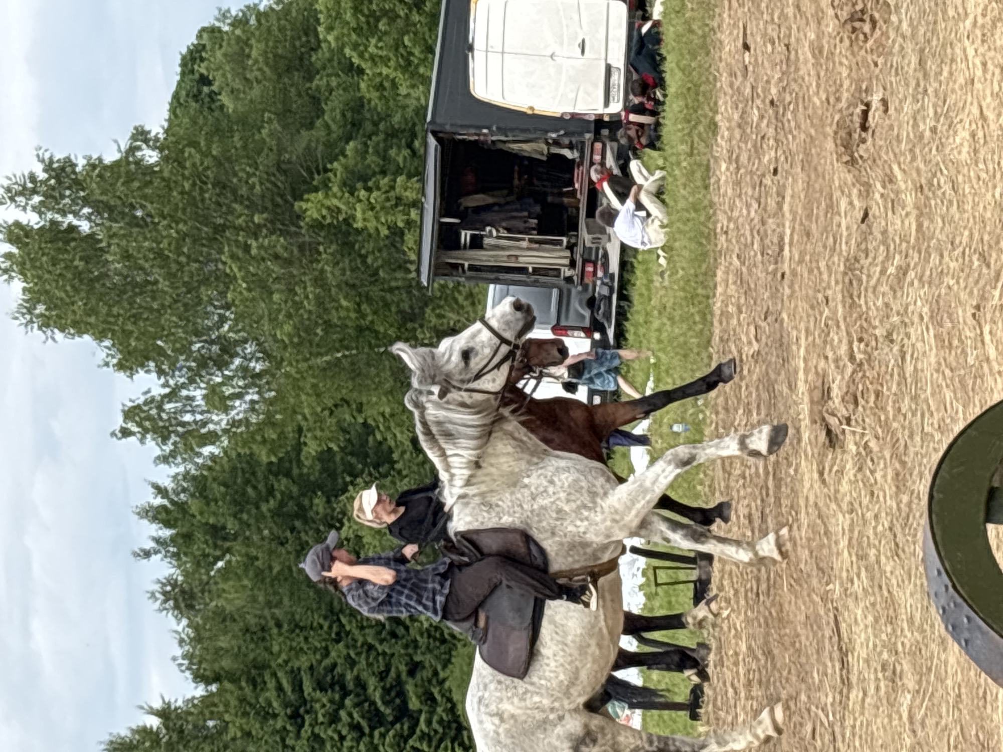In the city of Bornosovo, Russia, a grey speckled horse is ridden by a person wearing a dark and white plaid shirt, dark pants, and a baseball cap. Behind them, another person on horseback is visible. In the background, there is a black trailer with clothes hanging inside, and a white van. Several people are seated on the grass near the vehicles, some appearing to be resting or observing the activity. The scene is set outdoors with trees in the background and a ground covered in dry grass and straw.