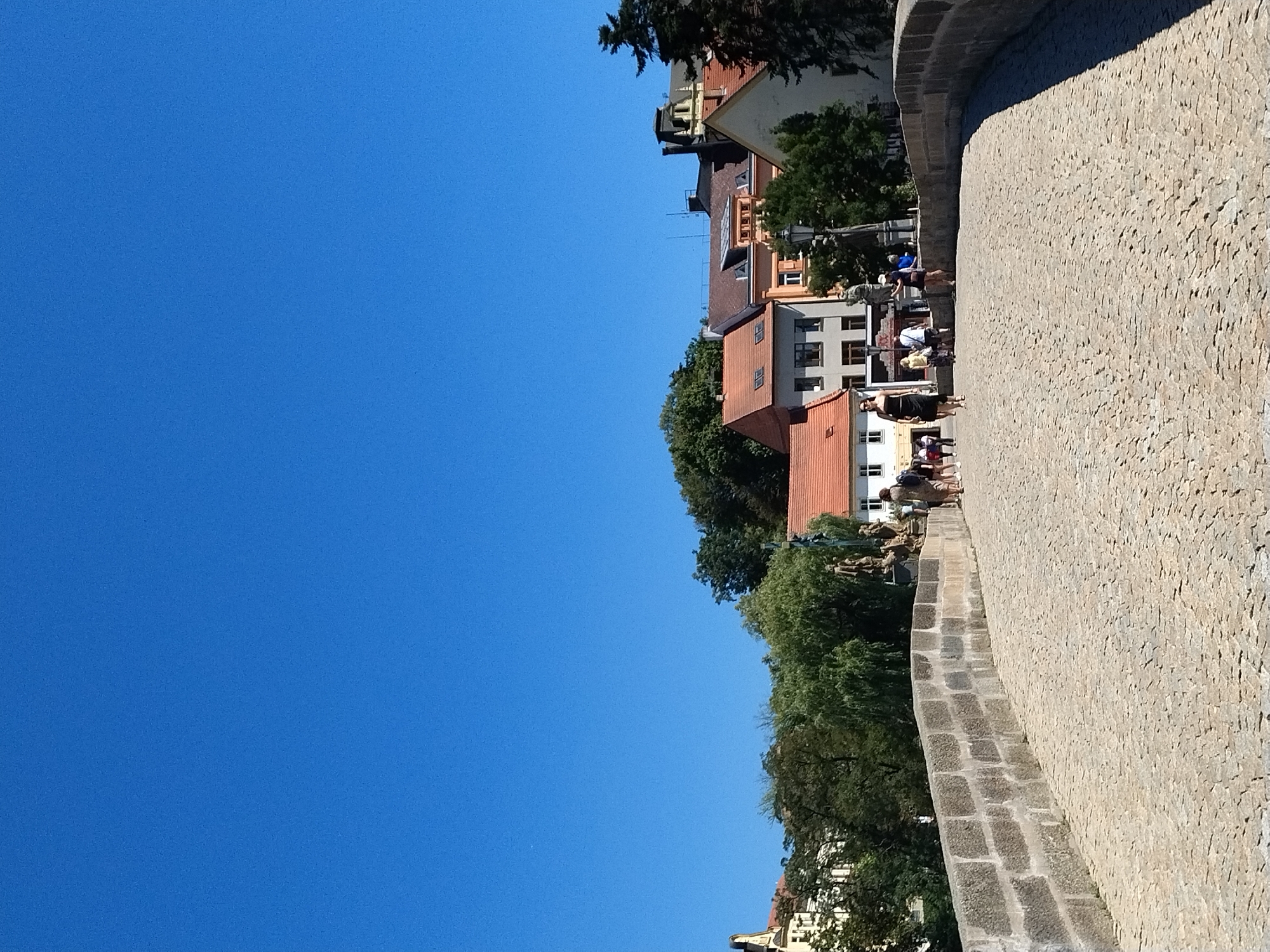 This image depicts a sunny day on the Stone Bridge in Písek, Czechia. A cobblestone pathway forms the foreground, leading up to and across the arched bridge. The bridge itself is constructed from large, grey stone blocks. Several individuals are visible on the bridge, walking in various directions. On the left side of the bridge, a woman in a black dress walks towards the camera. Further ahead, a group of people are gathered near a statue, with some pointing or looking. Buildings with tiled roofs and light-colored facades line the background, interspersed with lush green trees. The sky above is a clear, vibrant blue.