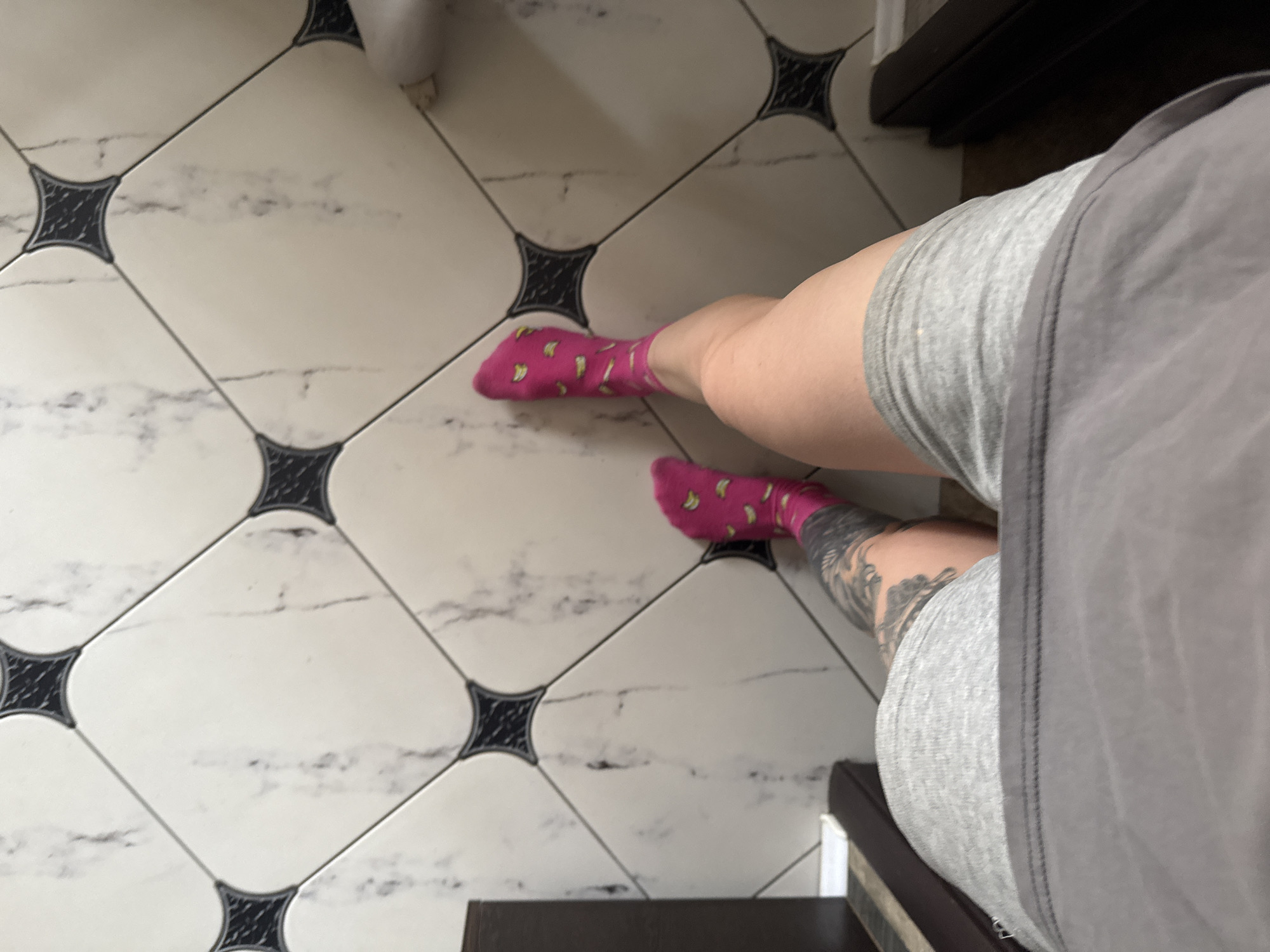 A top-down, slightly angled photograph shows a person's legs and part of their torso standing on a tiled floor. The floor tiles are square, white with black decorative elements at the corners, and have subtle marbling patterns. The person is wearing pink socks with yellow banana patterns. The person also appears to have a tattoo on their lower leg. They are wearing light grey shorts and a light grey shirt or dress. Part of a dark brown cabinet or furniture piece is visible on the right edge of the image. The photograph was taken in a bathroom. There is no information in the photo to indicate the location of Rostov-on-Don, Russia.