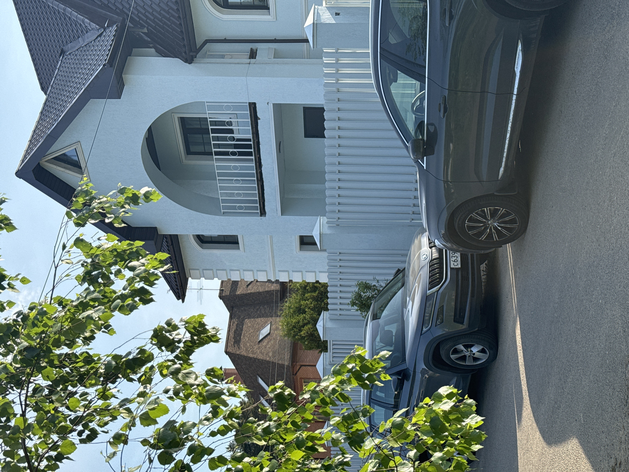 The image depicts a residential street in Селекционер-2 Снт, Russia. A light-colored, two-story house with a dark roof and arched balcony stands behind a white picket fence. Two dark-colored cars are parked on the street, with their front ends facing the viewer. A tree with green foliage partially obscures the view of the house and cars from the lower-left corner. The sky is visible in the upper-left corner and is clear. The house and cars are in focus, while the foliage and background elements are slightly blurred. The lighting suggests it is daytime.