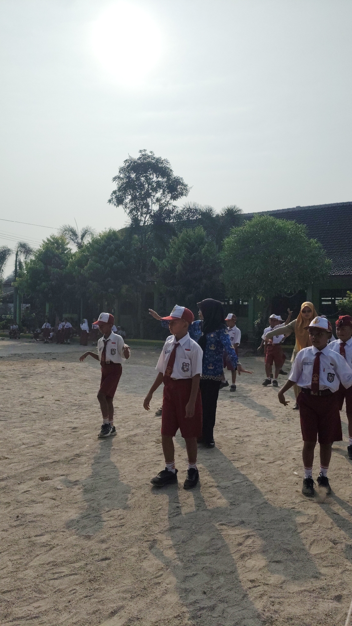 In Banjarsari, Indonesia, a group of elementary school students wearing red shorts, white shirts, and red and white caps are engaged in an outdoor physical activity on a sandy field. The students appear to be following instructions from two adult women. One woman, dressed in a blue patterned tunic and dark pants, stands behind a boy in the foreground, gesturing with her right hand. Another woman, wearing a yellow headscarf and light-colored clothing, is further back and also appears to be leading the activity. Trees and a building with a dark roof are visible in the background, under a bright, hazy sky. Shadows of the individuals are cast on the sandy ground, indicating it is daytime. Some other students are seated or standing in the background, to the left.