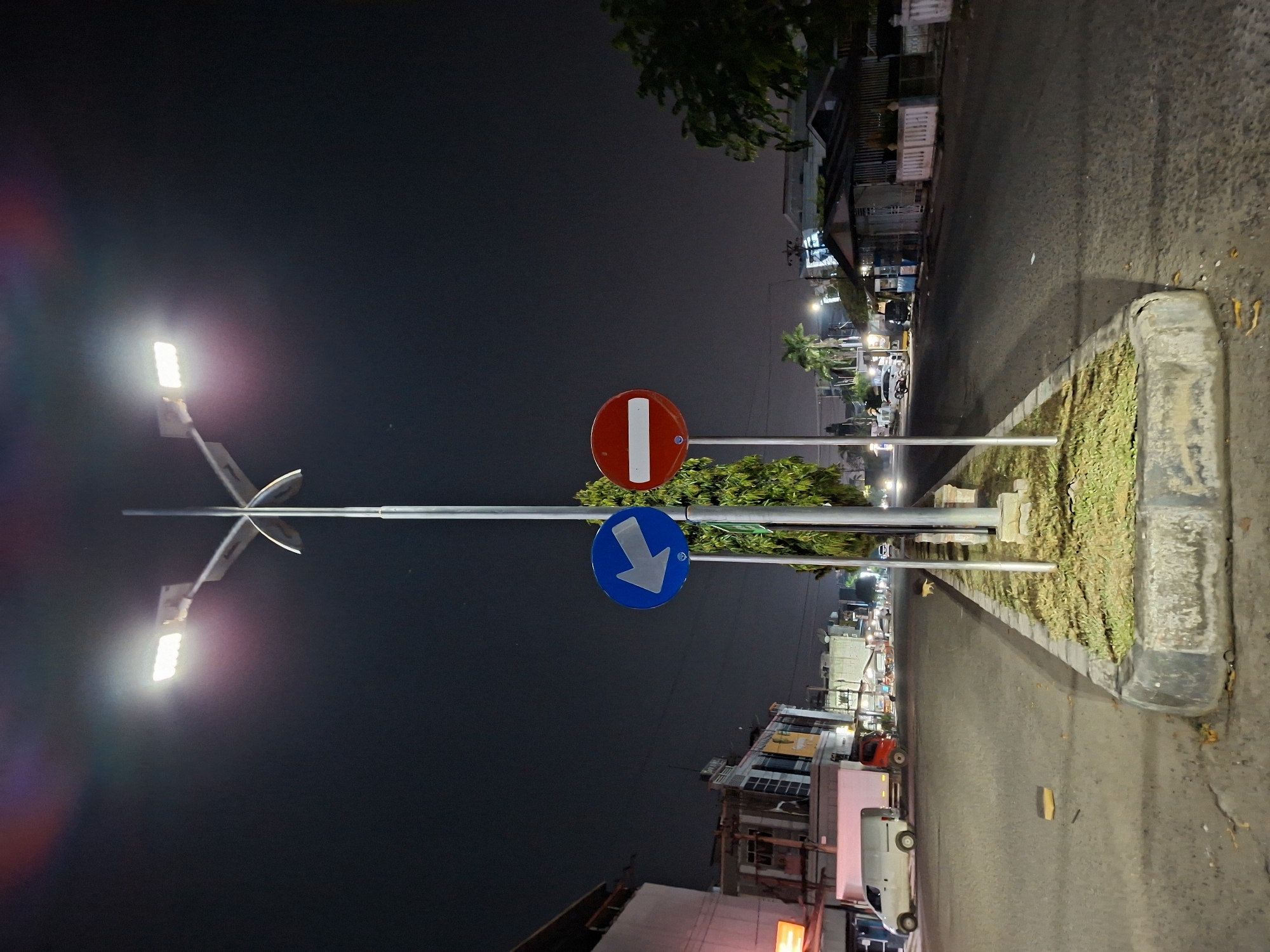 This is a night-time photograph of an urban street scene in Lhokseumawe, Indonesia. The image is oriented vertically but depicts a street running horizontally across the frame. The sky is dark, indicating it is night, and artificial lighting from streetlights and buildings illuminates the scene.
In the left foreground, a tall metal pole supports two bright LED streetlights. Mounted on the same pole are two circular traffic signs: a red "no entry" sign (a red circle with a white horizontal bar) positioned above a blue mandatory instruction sign (a blue circle with a white downward-pointing arrow). These signs suggest traffic control for vehicles approaching the vantage point of the camera.
To the right of the pole, a raised concrete median strip with short green grass separates the road. The asphalt road surface appears somewhat wet or reflective. In the background, on both sides of the street, are various buildings, some with illuminated windows or storefronts, indicating commercial or residential areas. A white van or car is visible on the right side of the road further down, and other indistinct vehicles can be seen in the distance, alongside more trees and foliage. No people are explicitly visible in the frame. The overall atmosphere is that of a quiet city street at night.