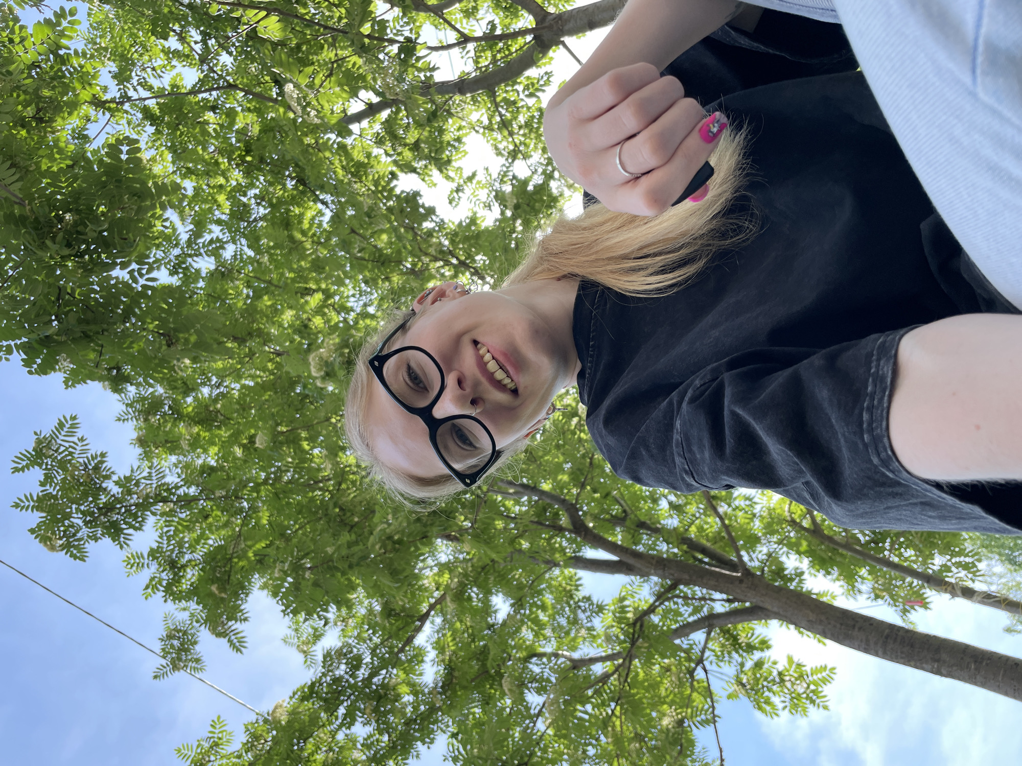 A low-angle, upward-facing shot depicts a fair-skinned woman with blonde hair, wearing black-framed glasses and a dark-colored shirt, against a backdrop of trees and a blue sky. The woman is smiling and looking directly at the camera. Her left hand is visible, displaying a silver ring and a pink nail with a design. The trees form a dense canopy above, partially obscuring the sky. The location, Izhevsk, Russia, is based on possible visual cues, but not discernible from the image alone. The specific details regarding activities, interactions, or precise geolocation are not present in the frame.