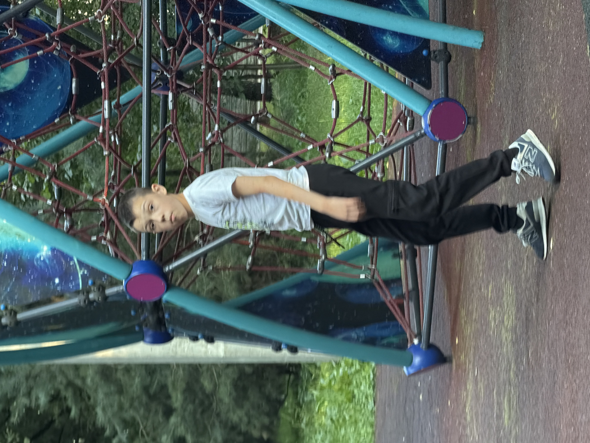 A young boy with short dark hair and a light-colored t-shirt and black pants is walking on a playground. He is looking at the camera with a surprised expression. The playground has a large, colorful climbing structure with blue and teal metal bars and red ropes. The ground is a reddish-brown rubber surface. Trees are visible in the background.