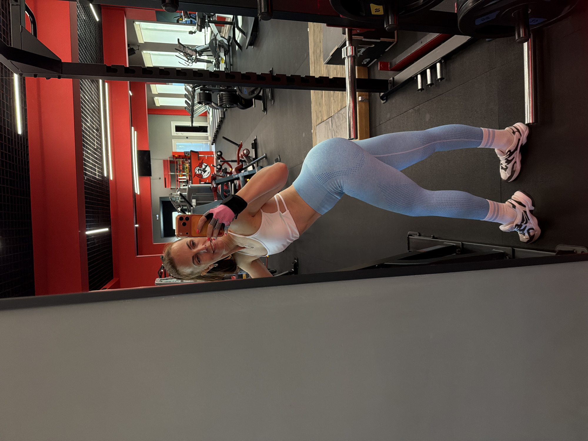 A young woman is depicted in a mirror selfie taken within a modern gym in Birobidzhan, Russia. She stands full-body, facing the mirror and holding an orange-cased smartphone to capture her reflection. She is smiling cheerfully at the camera.
The woman is dressed in athletic wear, including a white sports bra, form-fitting light blue seamless leggings with a dotted texture, white athletic socks with three dark stripes, and white and black athletic shoes. She also wears pink and black workout gloves on her hands.
The gym's interior design features red and black paneled walls, some adorned with horizontal strips of bright white LED lighting. The floor is dark, likely a rubberized gym surface. In the background, various pieces of exercise equipment are visible, including a black metal squat rack or power cage loaded with weight plates marked "15" and "20." Other indistinct gym machines and a red and white logo featuring a stylized fist are also present. The lighting is bright and artificial throughout the facility. The woman's pose and smile convey a sense of happiness and confidence.