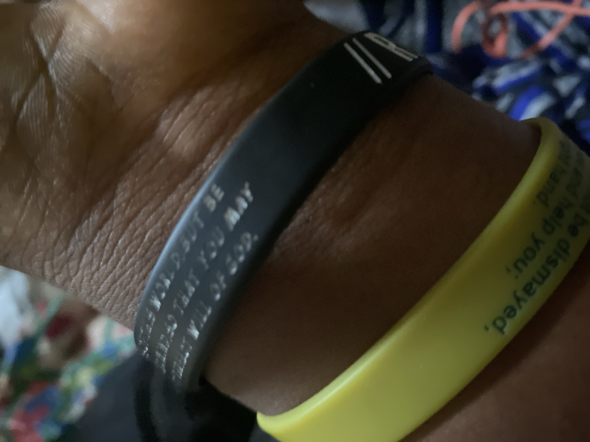 The image displays a close-up view of a person's wrist wearing two wristbands. The upper wristband is black and appears to have engraved text that is partially visible, reading "THIS WORD BUT BE READED THAT YOU MAY PERFECT WILL OF GOD." The lower wristband is yellow and also has engraved text, which is inverted and reads, "not be dismayed. not help you;". The background is blurred, showing a patterned fabric in shades of blue, white, and red, and a hint of skin texture on the wrist. There is no identifiable information about the location in Jauro Yinu, Nigeria.