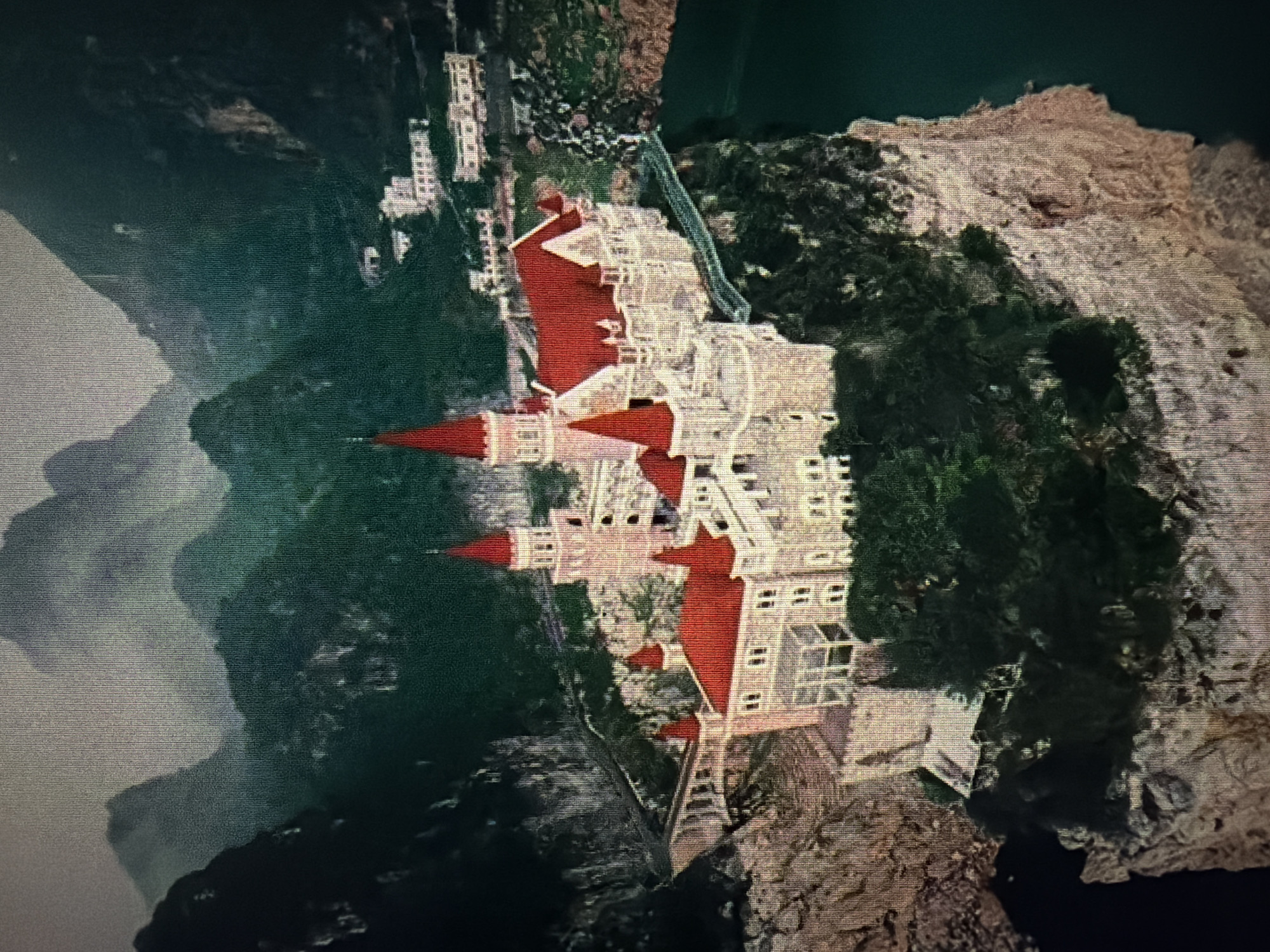 The image depicts a European-style castle with red roofs perched on a rocky cliff overlooking a body of water. The castle has multiple towers, crenellated walls, and numerous windows. The surrounding environment is mountainous and heavily forested, with a misty atmosphere. In the background, several modern buildings can be seen on a hillside. The image is a photograph, possibly taken from an aerial perspective, and appears to be of a fictional or reconstructed location, as there is no known castle matching this description in Baan Mae Nam, Thailand.