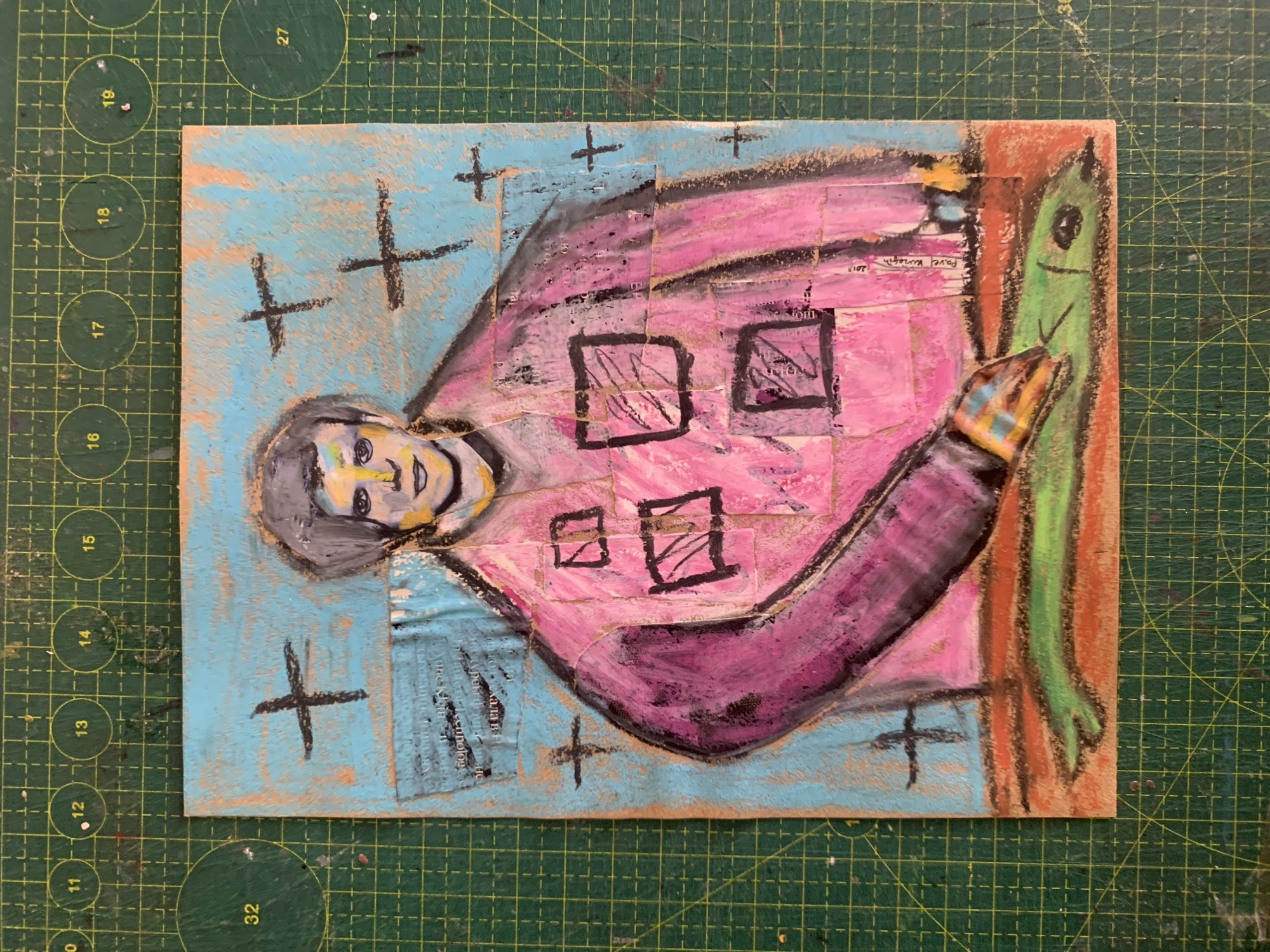 The image is a close-up, overhead shot of a mixed-media artwork on a green cutting mat. The artwork is a portrait of a person with dark hair, a yellow-toned face, and a pink garment. The background is a textured turquoise blue with black cross shapes. The person is shown from the chest up, with their left arm extended forward, holding a green object that resembles a fish. The artwork is pasted onto a textured brown surface. There are some collaged elements with text visible on the turquoise background. The bottom edge of the artwork shows a reddish-orange and green base. The cutting mat has a grid pattern with circles indicating measurements.
The provided image does not contain any information that could help identify the location in the city of Sergeevo, Russia.