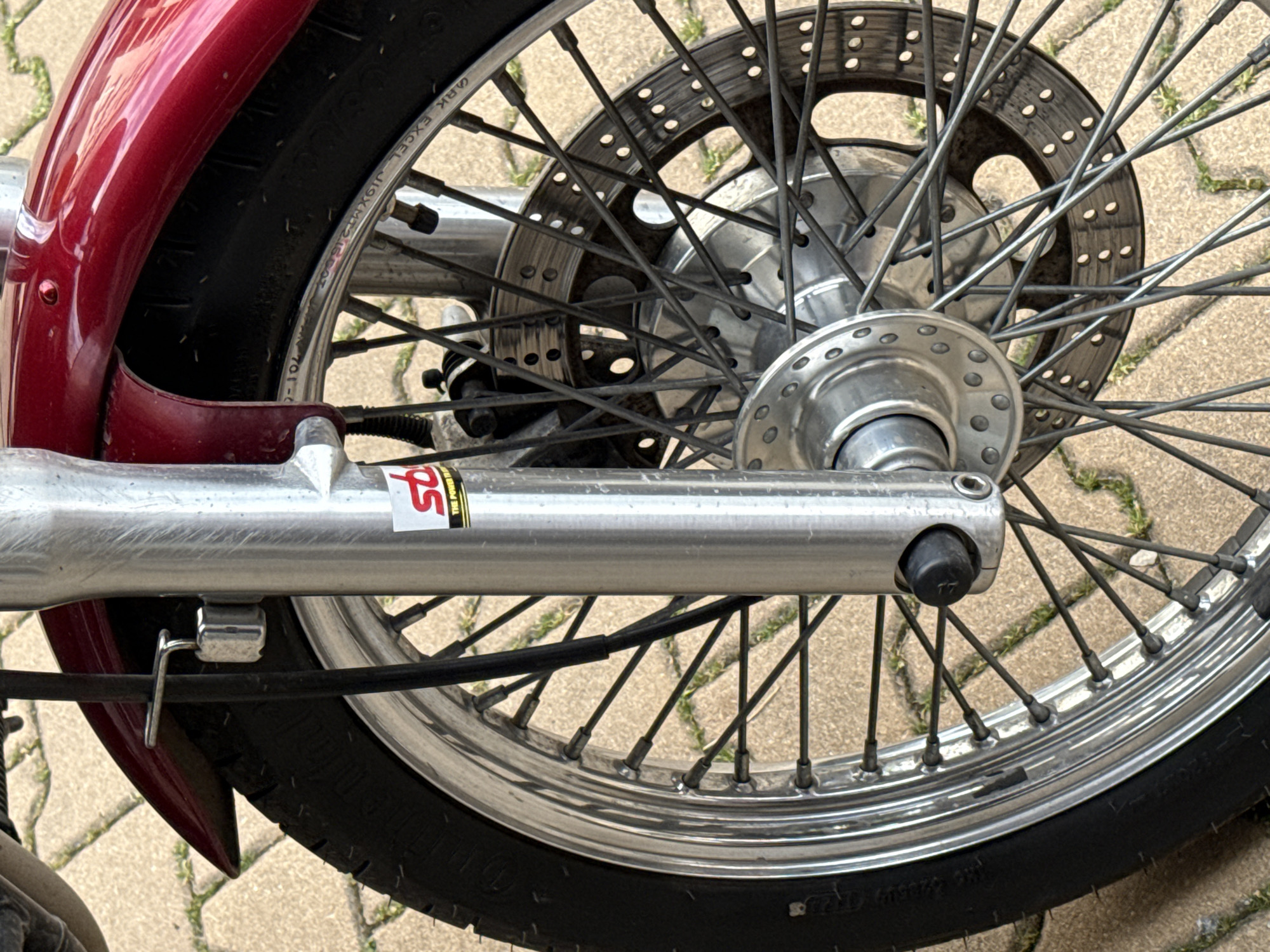 The image displays the front wheel assembly of a motorcycle, stationary outdoors on a paved surface in Sofia, Bulgaria. The dominant features include a dark red, glossy front fender positioned above a black rubber tire with visible tread. The wheel is a spoked type, featuring a chrome rim with manufacturer markings (partially visible "ARK EXCEL J19xMT2.15DOT") and numerous dark-colored spokes connecting to a central silver hub with a pattern of circular indentations. The front suspension consists of two silver-colored fork legs. A white sticker with red "sto" lettering and "THE POWER" text is affixed to the right fork leg. A black cable, consistent with a brake line or speedometer cable, runs vertically along the left fork leg, secured by a silver-toned metal bracket. A perforated silver disc brake rotor is visible on the right side of the wheel, positioned behind the spokes. The ground beneath the motorcycle is composed of light brown/beige rectangular pavers, with green vegetation, likely moss or weeds, growing within the cracks. The scene is illuminated by natural daylight; no individuals or active events are depicted.