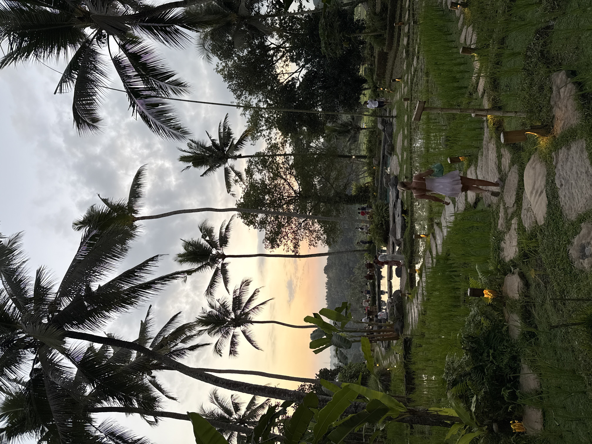 A woman in a white dress walks away from the camera on a stone pathway through a rice paddy at dusk in Ubud, Indonesia. Palm trees are visible in the foreground, and the sky is a mix of grey clouds and a soft orange sunset. Several people are gathered in the background near a body of water, and small lights illuminate the pathway. The scene depicts a serene, tropical evening in a rice-growing region.