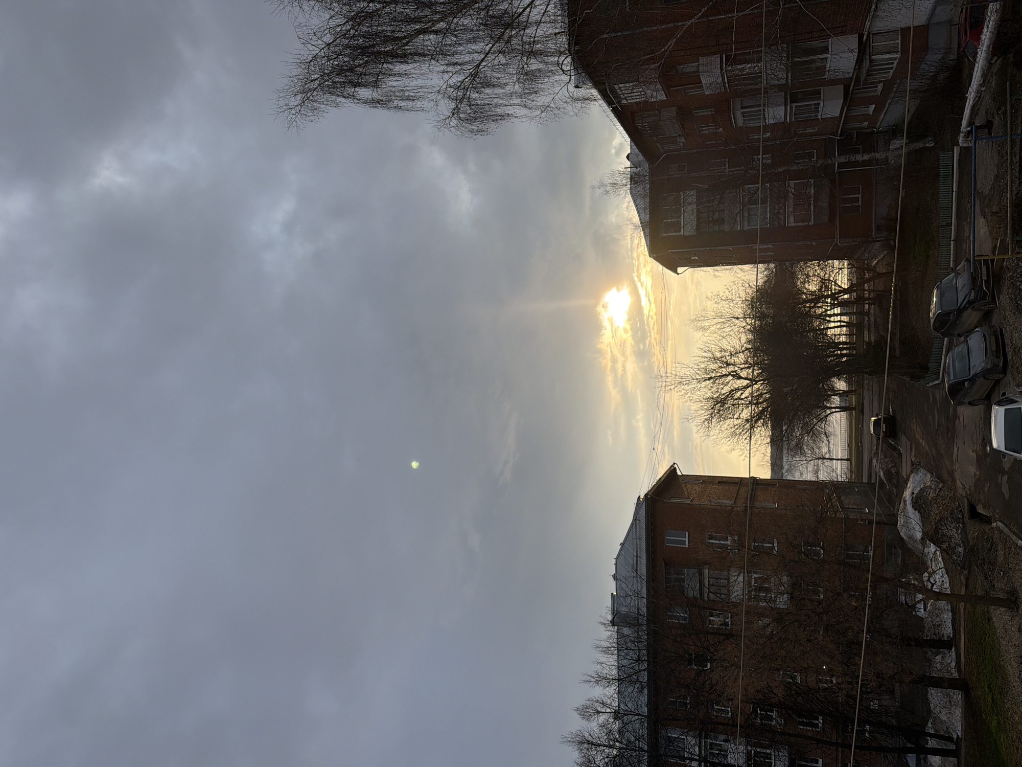 The image depicts an urban scene in Votkinsk, Russia, at sunset on an overcast day. Two brick apartment buildings frame a street with parked cars and bare trees. The sun is visible through a break in the clouds, casting a bright glow over the horizon. The overall scene suggests a cold, possibly winter, day in a residential area. There are no people visible.