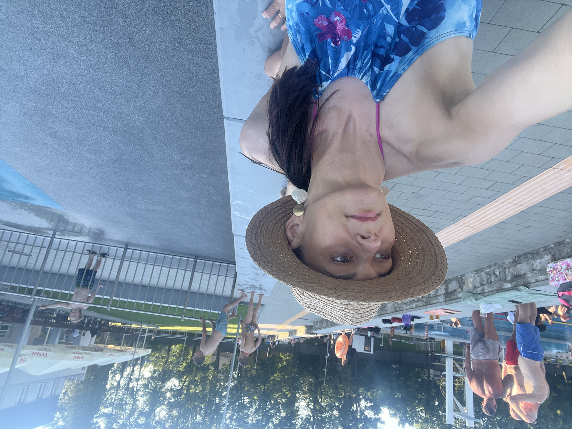 In an outdoor swimming pool area, a woman wearing a straw hat and a blue floral swimsuit is in the foreground, looking to her left. She has her hair in a ponytail and is wearing large, circular earrings. Behind her, several people are visible. In the background, there is a diving platform, trees, and umbrellas from a refreshment area. The ground is paved with gray tiles and there is a pool with blue water. Some children are playing near a metal fence.