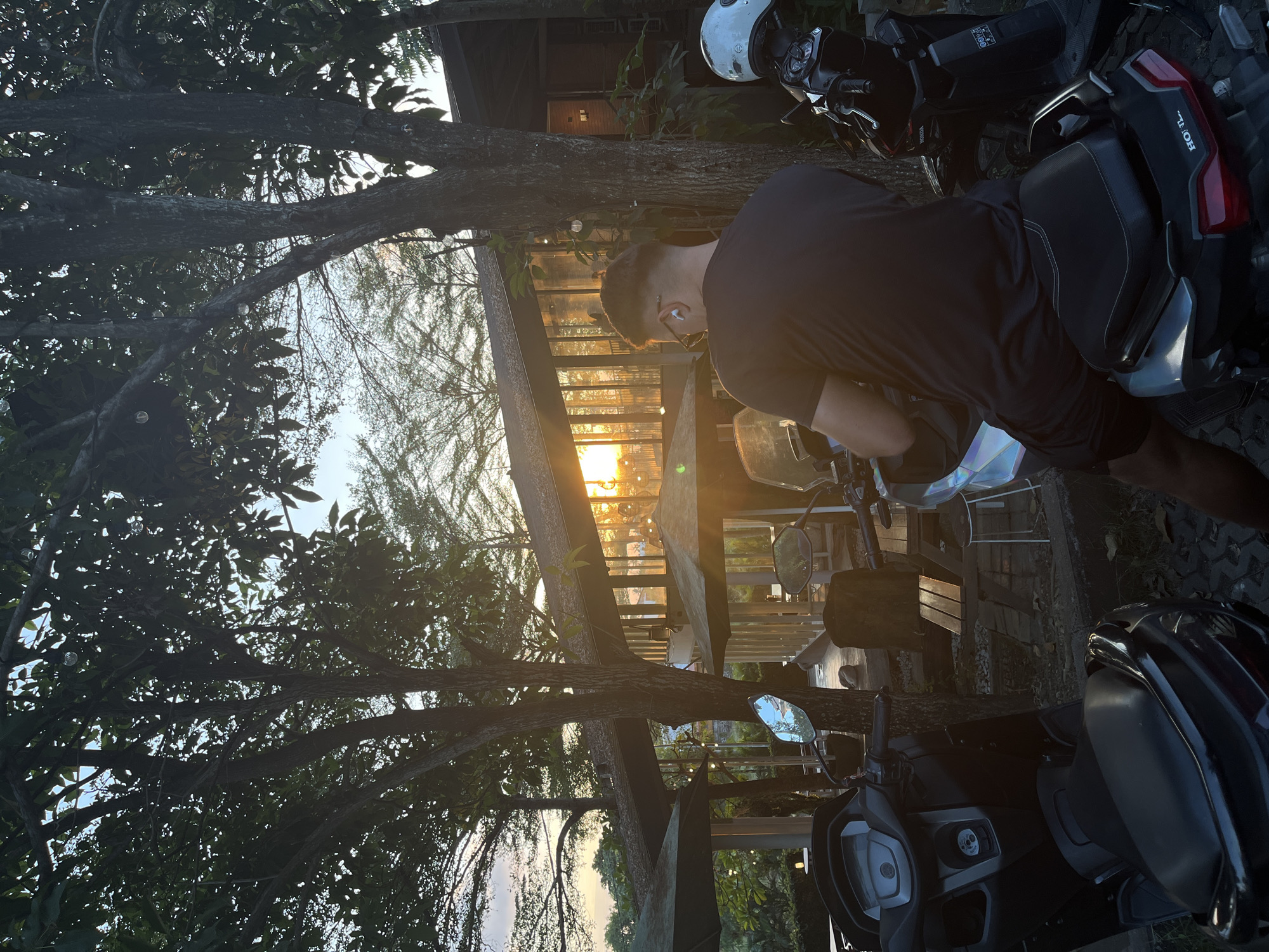 A back view of a man sitting on a scooter in the foreground, facing a building with large windows reflecting the sunset. The man is wearing a black t-shirt and black shorts, with earbuds in. Two scooters are parked next to him. In the background, there are trees with lush green leaves and outdoor seating areas with umbrellas. The lighting suggests it is late afternoon or early evening. The scene appears to be an outdoor cafe or restaurant in North Kuta, Indonesia.