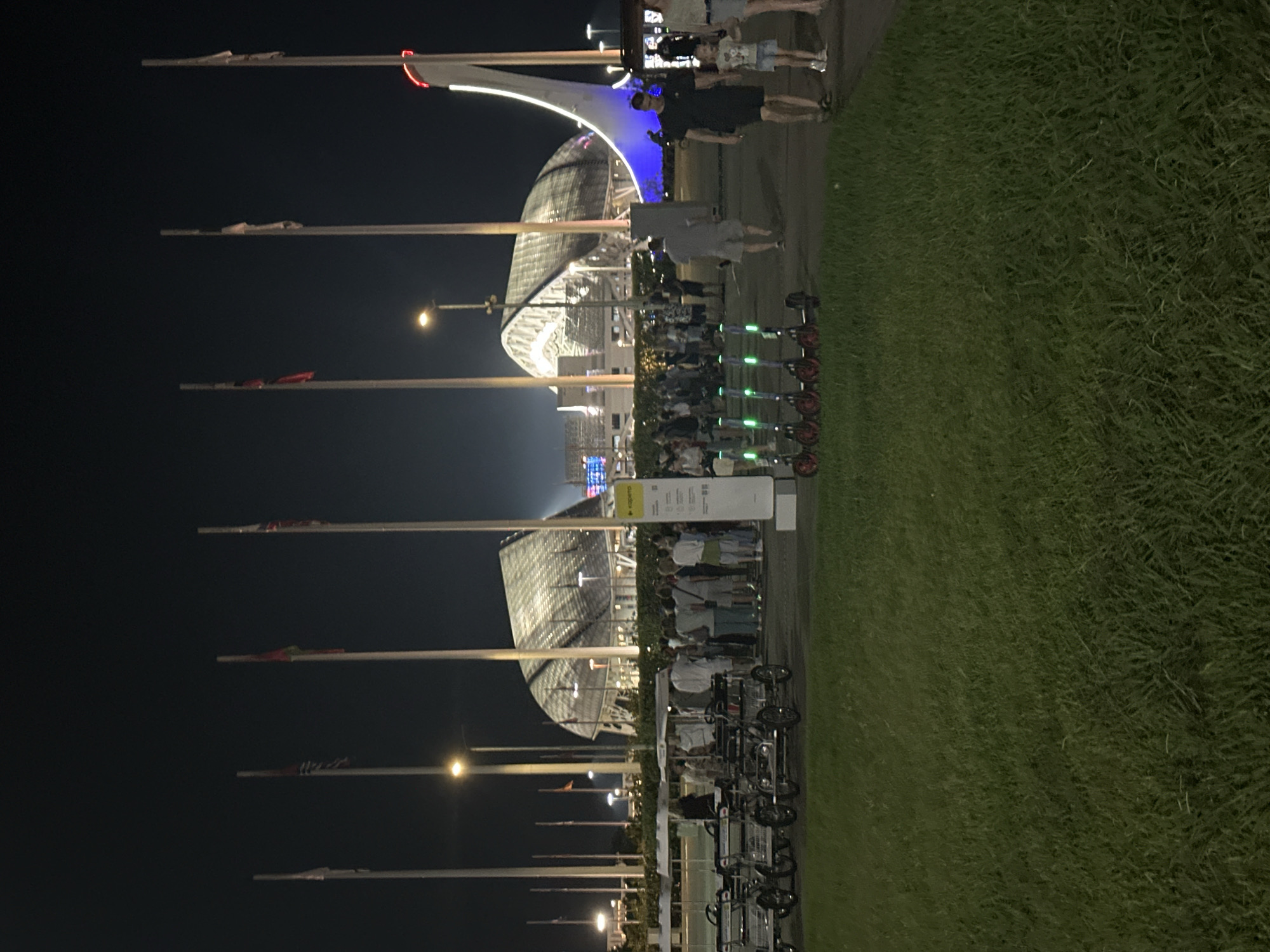 The image depicts the Fisht Olympic Stadium in Sochi, Russia, at night. Several tall flagpoles with flags are positioned in front of the stadium, which features a unique, translucent roof illuminated with lights. A large crowd of people is gathered in the foreground, appearing to be observing the stadium or waiting. In front of the crowd, there are several pedal-powered carts or "quadricycles" lined up. To the right of the carts, a row of electric scooters is parked. People are walking around the area, some in casual attire. The ground in the immediate foreground is covered in grass, while the area behind the people and carts appears to be paved. The overall scene suggests a public gathering or event at a prominent landmark in Sochi.