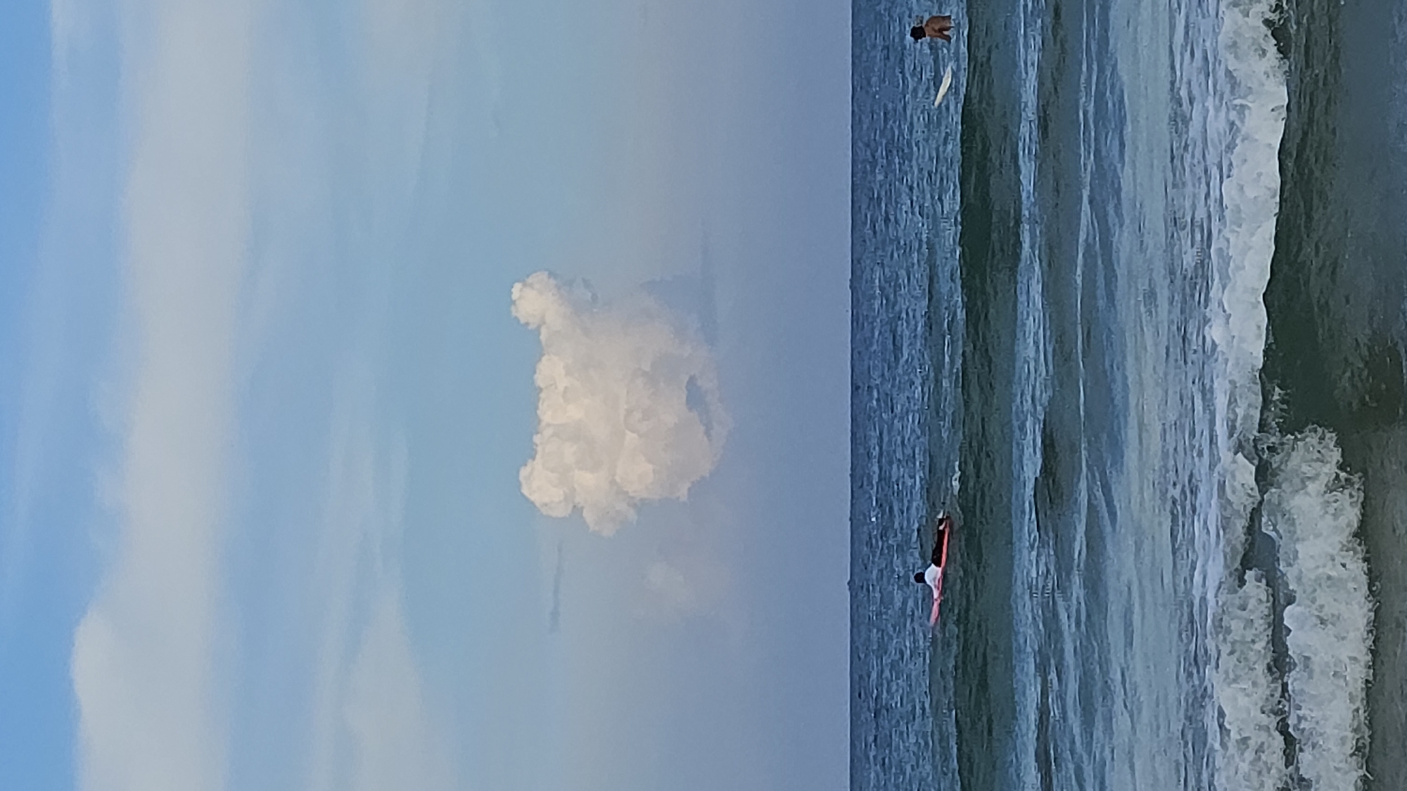 The scene is a vertically oriented image of the sea and sky, likely taken in Cam Lam, Vietnam. The sky, occupying the left side of the frame, is light blue with soft white clouds. A prominent cumulus cloud is centered in the frame. The sea extends across the right side of the image, transitioning from a darker blue in the distance to a lighter, greenish-blue near the shore. Two individuals are visible in the water. One person is farther out, floating near a white object (possibly a surfboard). A second person is closer to the shore on a red object (likely a surfboard), appearing to be paddling. White waves are breaking near the shore.