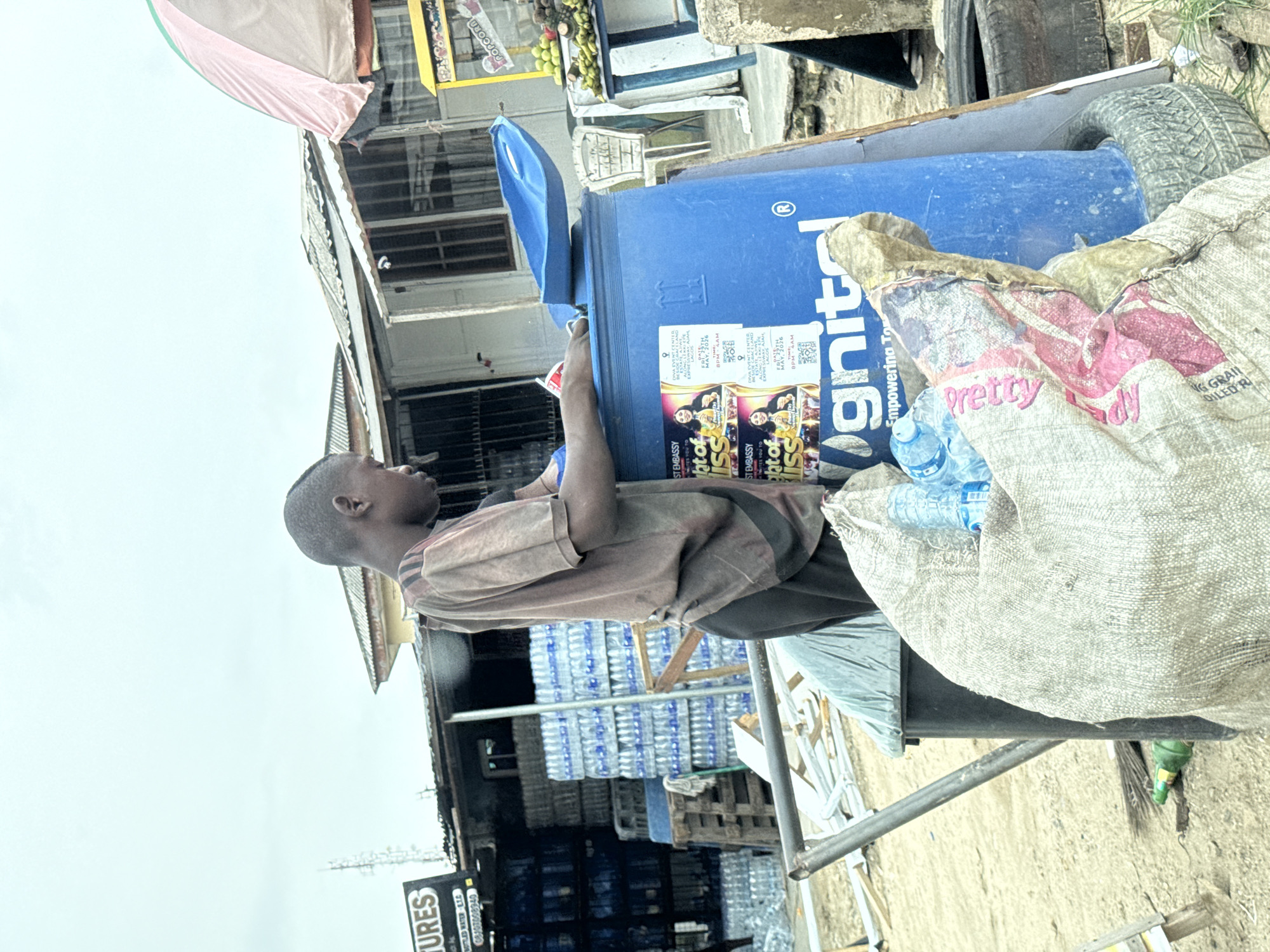 A young boy is standing beside a blue plastic trash bin filled with plastic bottles and other trash, in Lagos, Nigeria. The trash bin has the word "Ignite" printed on its side, and flyers for an event called "Light of Bliss" are also stuck to the bin. There are stacks of bottled water in the background, on a platform. A large white sack with the word "Pretty" printed on it is also near the bin. The boy is next to a cart.