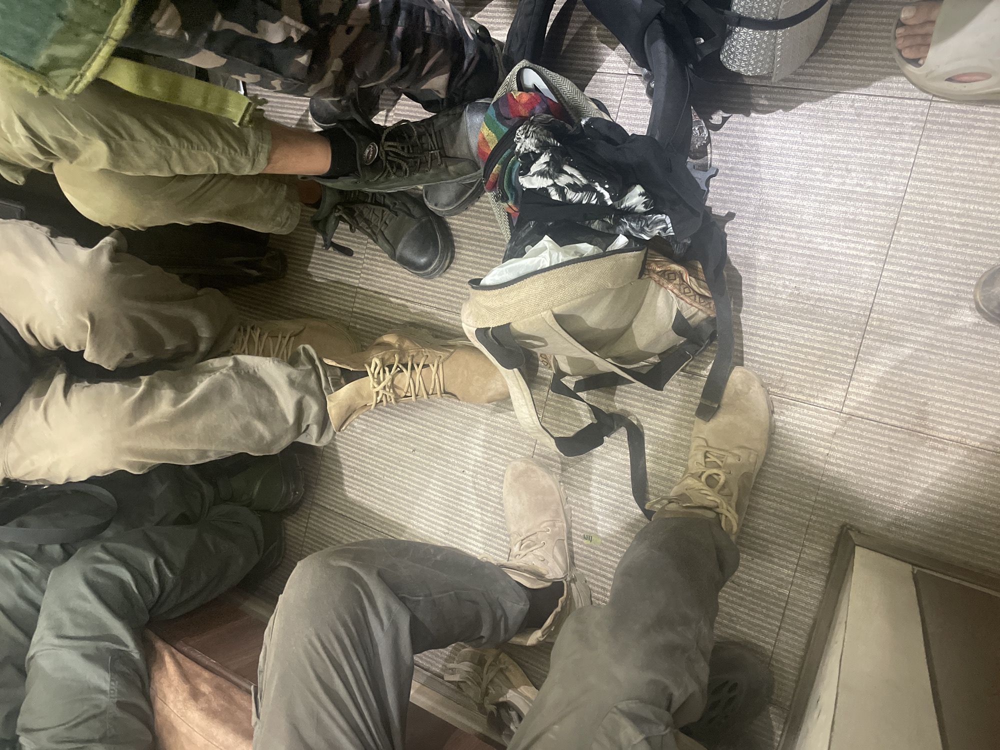 The image shows a close-up, overhead view of multiple people seated on a patterned floor. Five sets of legs and feet are visible, dressed in various pants and boots. The pants include camouflage, green, khaki, and gray materials. The footwear consists of several pairs of boots, including tan, gray, and dark green styles. There is also a bare foot wearing a sandal.
Between the legs, there are backpacks and other bags, including one open bag with what appears to be clothing inside. The setting is indoors with a tiled or carpeted floor.
The specific location of Mira-Bhayander, India is not evident from the details in the image. Therefore, I cannot pinpoint an exact location within the city.