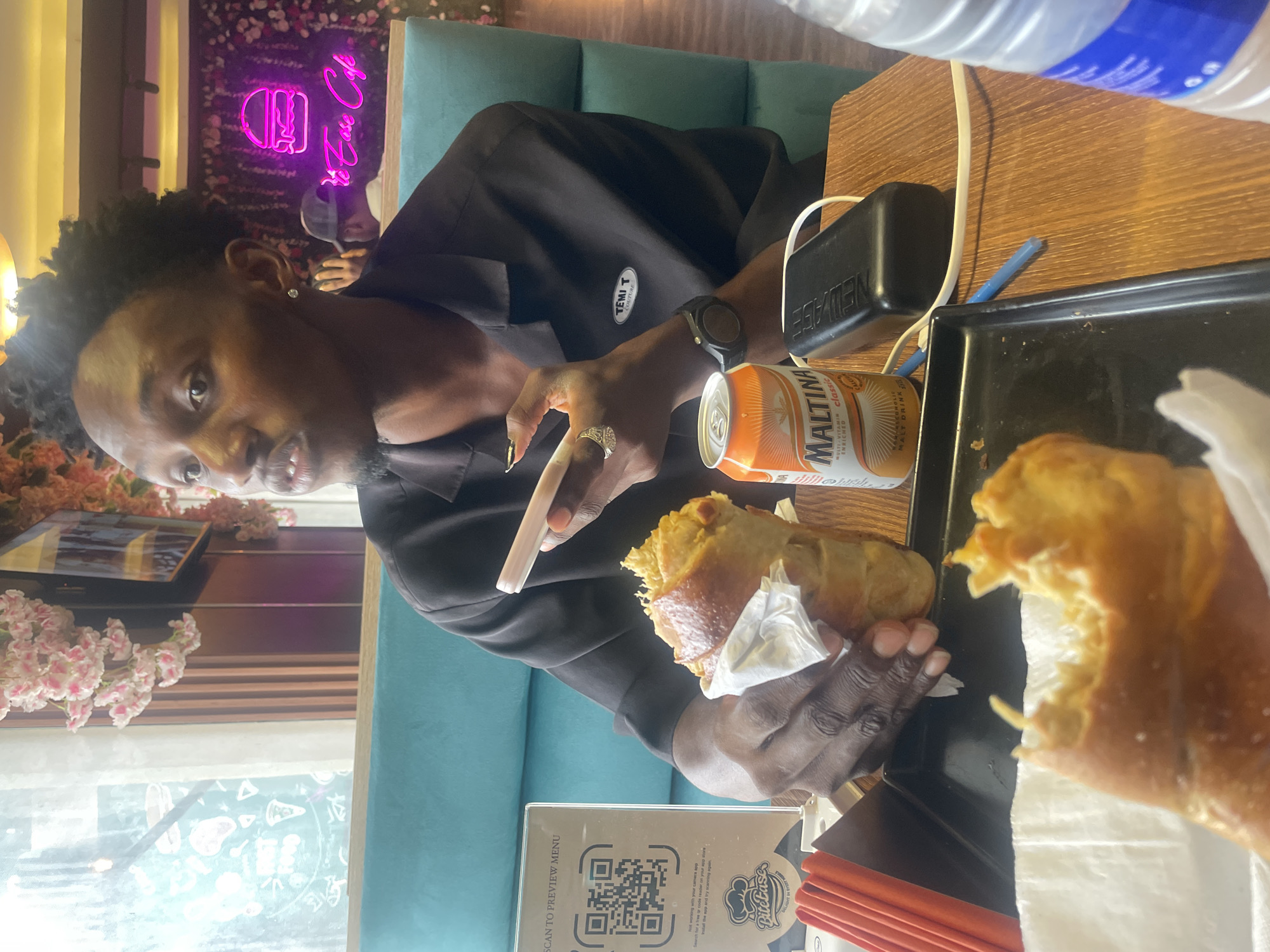 A person is seated at a table in a restaurant or cafe in what appears to be Adankolo, Nigeria. The individual is dark-skinned and wearing a black shirt. They are holding a smartphone in one hand and a partially eaten sandwich wrapped in paper in the other. An open can of Maltina, a popular malt drink in Nigeria, is on the table next to a black object, possibly a power bank. A blue pen lies nearby. A black serving tray contains another piece of the same sandwich, partially obscured by the wrapper. A QR code menu from "Bitetaste" is also on the table. The environment features artificial floral arrangements and a neon sign that reads "Ve Ease Cafe." A teal-colored cushioned bench seat is visible. A bottle of water is present in the upper right corner.