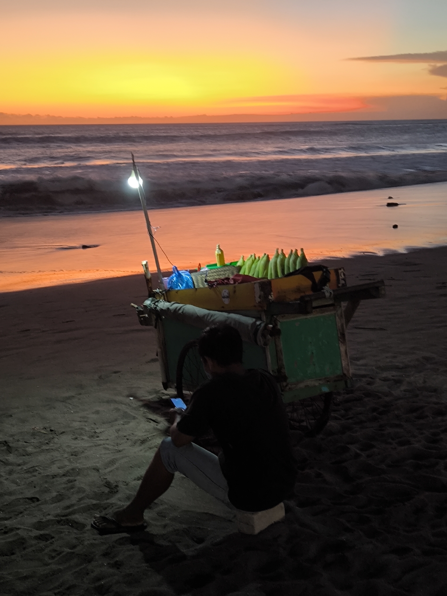 A vendor is seated on the beach in North Kuta, Indonesia, using his phone next to a green pushcart. The cart contains wrapped ears of corn, drinks, and a small light source. The sun is setting over the ocean in the background.
