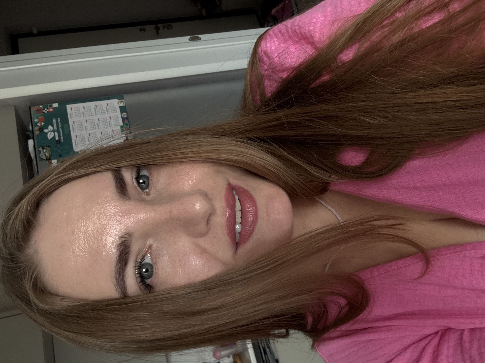 A woman with long brown hair and blue eyes is shown in a close-up selfie. She is wearing a bright pink top. Her lips are painted with a reddish-pink lipstick, and she has a small, pearl-like embellishment on one of her teeth. Her skin appears somewhat shiny, suggesting it might be humid or she is wearing illuminating makeup.
In the background, a refrigerator is visible, adorned with a calendar. The calendar is green and features various images and text, including what appears to be a grid for each month of the year, suggesting it's a planning tool. The overall environment seems to be indoors, possibly a kitchen or a similar domestic setting. The lighting suggests it is daytime, with light coming from an unseen source that casts highlights on her forehead and cheeks. There are no discernible signs indicating the location is specifically within the city of Kazan, Russia.