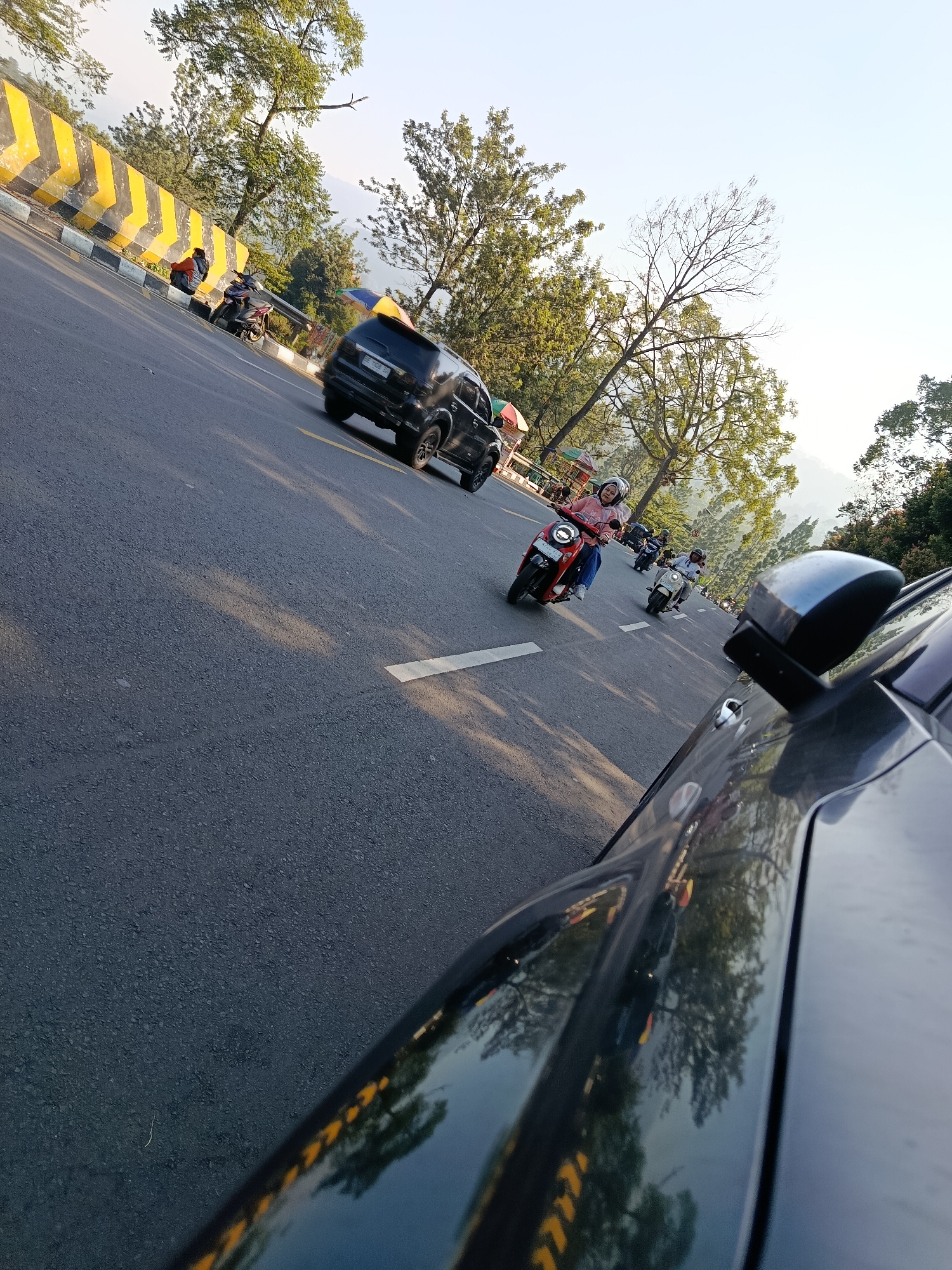 This image captures a street scene in Cisarua, Indonesia, with a focus on traffic and the surrounding environment. The road is paved and appears to be a two-lane highway with distinct yellow and black chevron markings on a concrete barrier on the left side. The time of day appears to be late afternoon or early morning, indicated by the long shadows cast on the road and the warm lighting.
Several vehicles are present, including a black SUV driving in the left lane and multiple motorcycles in the right lane. One motorcycle in the foreground is red and being ridden by a person wearing a pink shirt and a white helmet. Another motorcycle, white in color, is also visible with a rider. The motorcycles are in motion, suggesting a flow of traffic.
The environment is characterized by lush greenery and trees lining the road, indicating a semi-rural or hillside location within Cisarua. In the background, a hazy mountainous landscape is visible, a common feature of the Cisarua region known for its natural beauty. The sky is clear and light blue, with the sun's position low on the horizon, contributing to the lighting conditions.
The perspective of the photo is from inside a vehicle, looking forward and slightly to the side, with the reflection of trees and traffic visible on the vehicle's surface. This viewpoint adds a sense of immersion to the scene.
