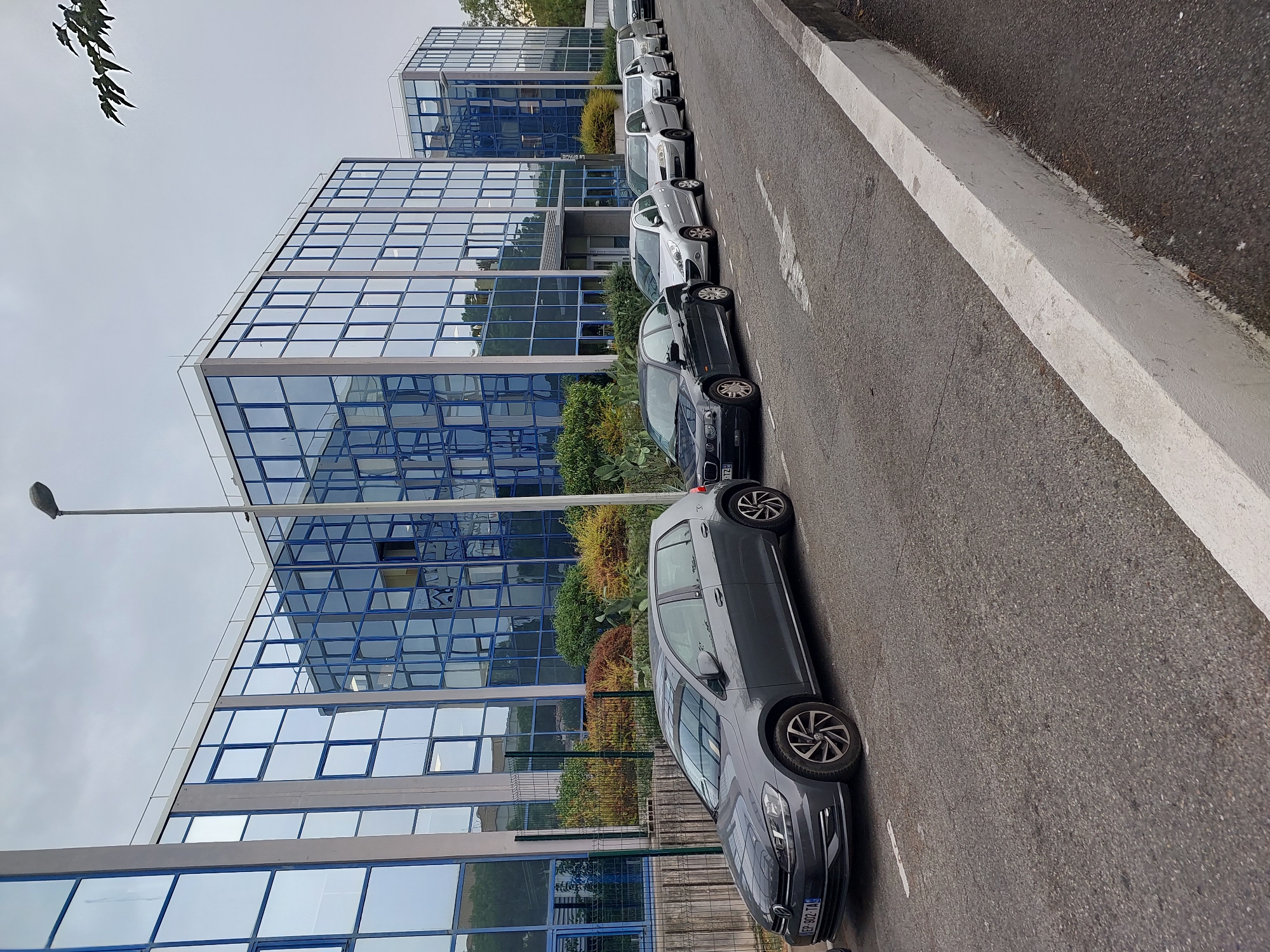 The image shows a street in Nice, France, lined with cars parked in front of a modern office building. The building has a blue glass facade and appears to be several stories high. A street lamp stands on the sidewalk next to the building. The cars are parked in a row along the street, with the closest car being a dark grey Volkswagen Golf. The sky is overcast, and the overall scene suggests a typical urban environment. The image is taken from a slightly elevated angle, looking down the street.