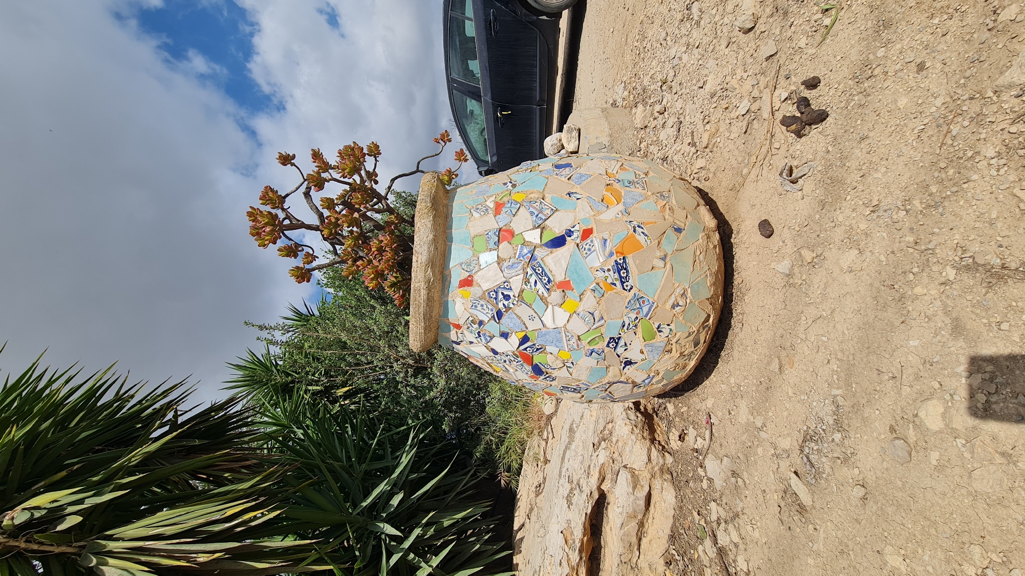 The image depicts a large, mosaic-covered pot filled with greenery, situated outdoors in the Lachish Regional Council, Israel. The pot features irregular fragments of tile in blue, white, orange, and yellow adhered to its surface. A succulent plant with reddish-brown leaf clusters grows from the pot. Behind the pot, dense foliage includes spiky yucca plants and other shrubbery. Above the pot, a partial view of a black vehicle is visible against a sky with scattered clouds. The ground around the pot is dry and rocky, with small, dark droppings scattered nearby. The image is oriented sideways.