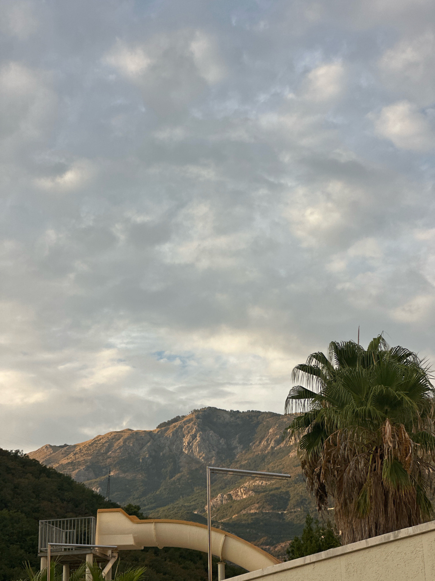 The image captures a scene in Boreti, Montenegro, with a dramatic mountainous backdrop. In the foreground, a water slide structure with a cream-colored slide and metal railings is partially visible on the left. To the right, a large, lush palm tree with drooping fronds dominates the view, its base partially obscured by a light-colored wall. The middle ground features a densely forested hillside leading up to rugged, sun-drenched mountains. The sky above is filled with a complex pattern of grey and white clouds, with some patches of lighter sky peeking through, suggesting it might be overcast or nearing dusk. The overall impression is one of a resort or outdoor recreational area situated in a scenic, mountainous coastal region.