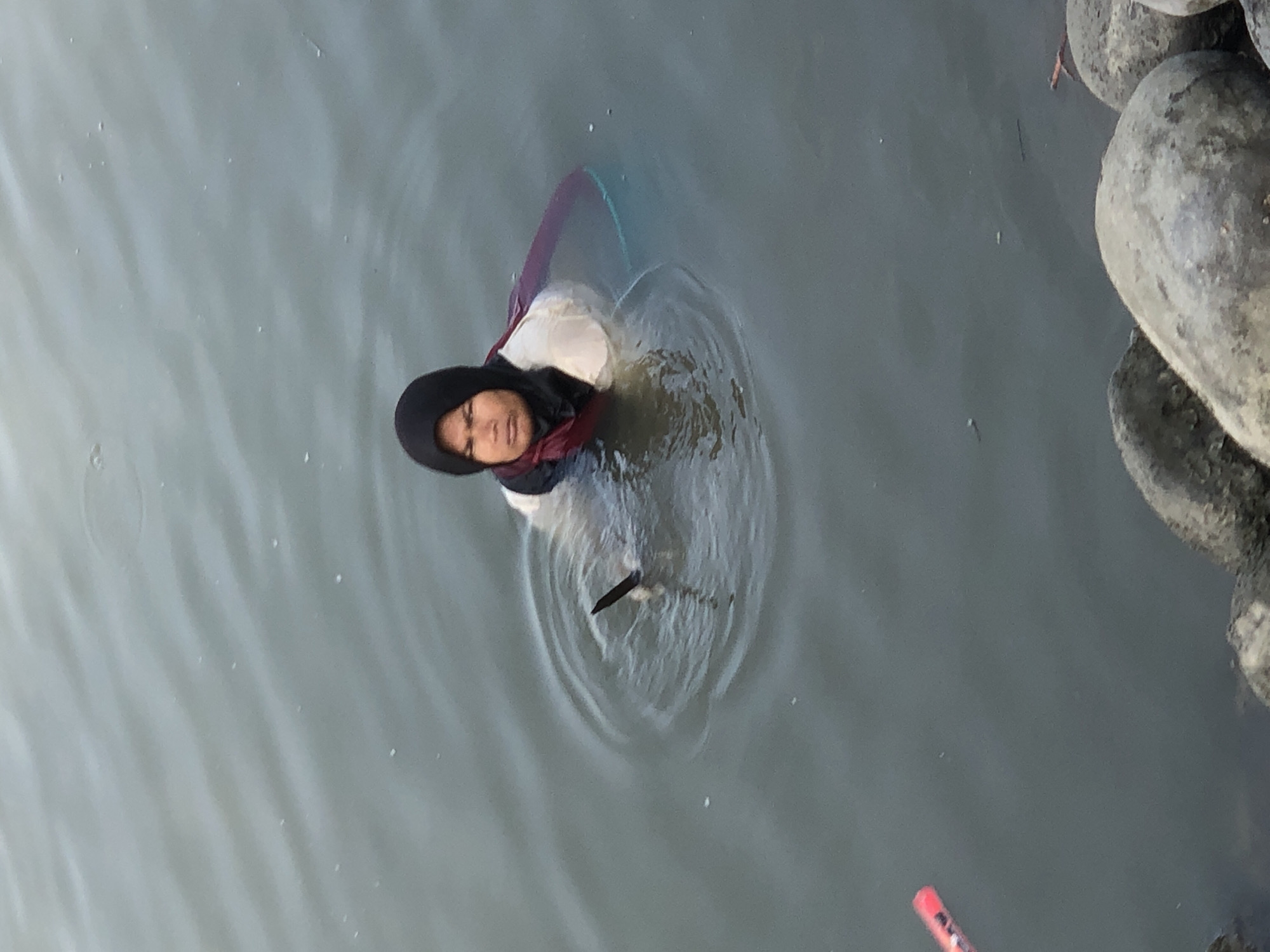 A person wearing a dark hijab and a light-colored top is partially submerged in murky water, with their upper body visible. They appear to be in shallow water near a rocky shore. A dark, pointed object, possibly a fishing spear, is held in their right hand, submerged in the water. The person's facial expression is neutral. In the background, the water is calm with ripples around the person. A red object, possibly a marker or buoy, is visible in the lower left corner of the image. The overall scene depicts a person engaged in an activity in a body of water, possibly fishing or collecting something from the water, in Lhokseumawe, Indonesia.