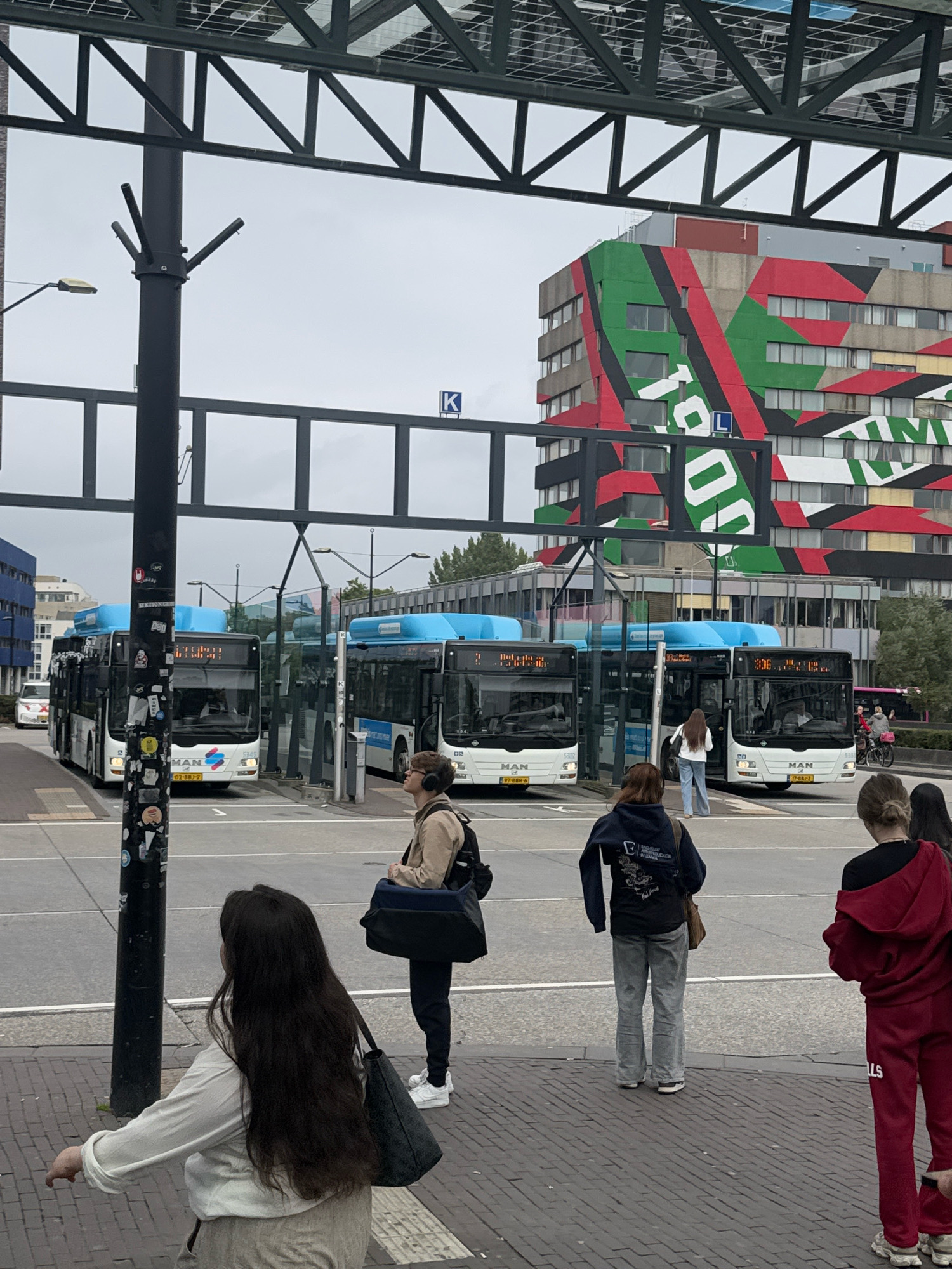 The image depicts a street scene in Nijmegen, Netherlands, specifically at the bus station located near the central train station. In the foreground, four individuals are visible, three of whom are facing away from the camera. One person, wearing a white top and light-colored pants, is turned to their left and appears to be walking away. Another person, with long dark hair, is also facing away, holding a dark bag. A third person, wearing a dark blue hoodie and grey pants, stands looking towards the buses. The fourth person, in the background and to the right, is partially obscured and wears headphones and a light-colored jacket.
Several buses, predominantly white with blue roofs and destination displays, are parked at the bus stop. The buses have visible license plates and company branding, including "MAN". Behind the buses, a distinctive building with a colorful, abstract geometric facade in red, green, and black dominates the background, identifying the location as Nijmegen. The environment appears to be an urban setting with a covered bus shelter structure overhead, supported by metal poles. The sky is overcast. The ground is paved with brickwork. A street light is visible on the left side of the image.