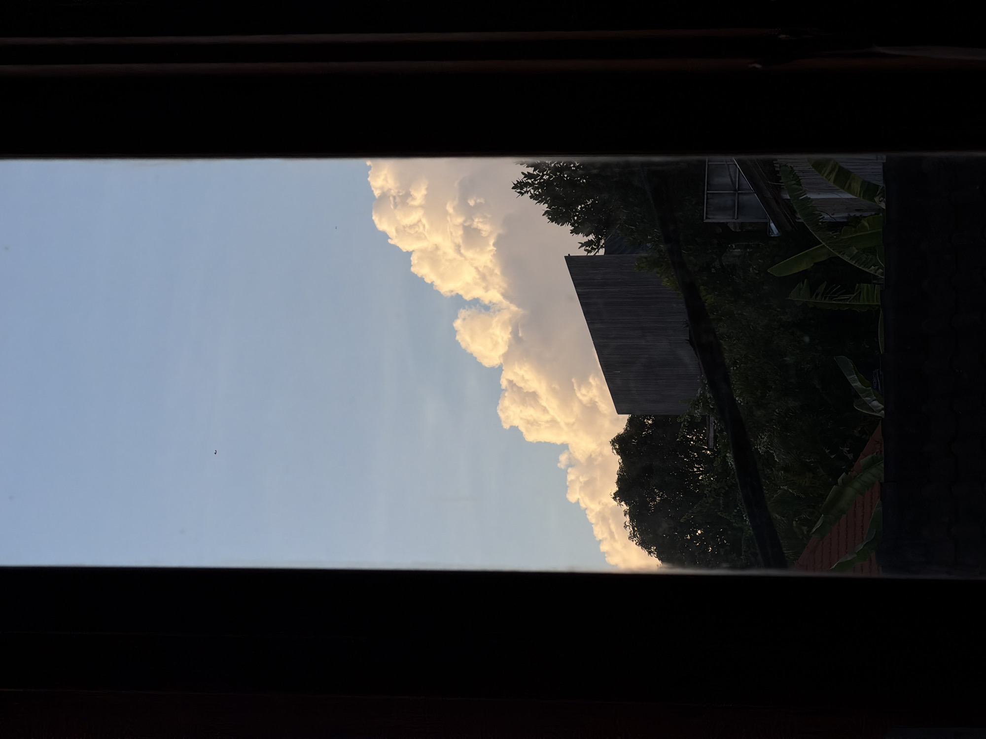 The image shows a view from inside a building in North Kuta, Indonesia, looking out through a window. The window frame is dark and creates a border around the scene outside. The sky is visible, with a gradient from light blue to a brighter, whiter hue, and there are large, fluffy cumulus clouds, some with a golden glow, indicating either sunrise or sunset. A building is visible adjacent to a dark green treeline. A small dark spec that could be a bird is visible in the upper left of the open window pane.