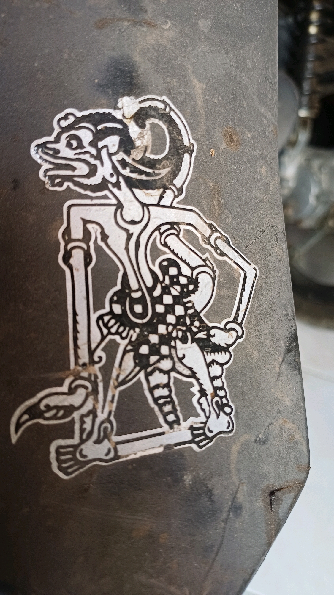 The image is a close-up, eye-level shot of a sticker depicting a Wayang Kulit puppet on a textured, dark gray, possibly metal surface. The sticker itself is black and white, featuring a character with an elongated head, sharp teeth, and a distinct headdress. The puppet has thin, jointed limbs and is adorned with a checkered pattern on its torso. The surface the sticker is on shows signs of wear and tear, with visible scratches and dirt specks. The background is blurred, with hints of metallic objects and a light-colored surface, suggesting an outdoor or workshop environment. The specific location in Wonokerso, Indonesia, cannot be determined from the visual information provided in the image.
