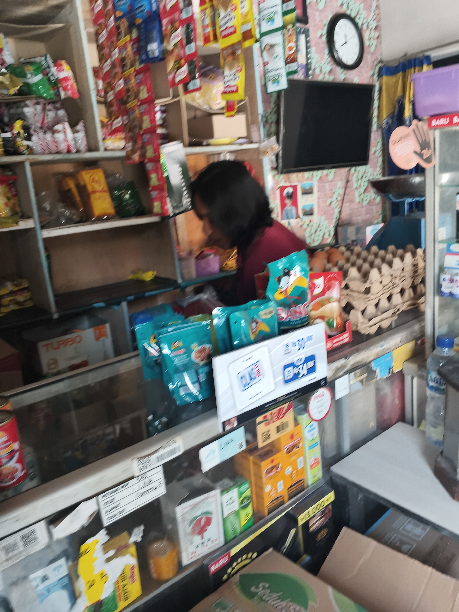 The image depicts the interior of a small convenience store, likely a "warung" or local shop, in Bandar Lampung, Indonesia. Shelves are stocked with various packaged goods, including snacks, drinks, and household items. In the background, a person with dark hair is partially visible, seemingly attending to the store's inventory or serving a customer. Prominent items include cartons of eggs, bottles of water, and colorful packaging of food products. A wall clock with Roman numerals is visible on the upper right, and a small television screen is mounted on the wall. The overall impression is that of a typical, cluttered neighborhood store.