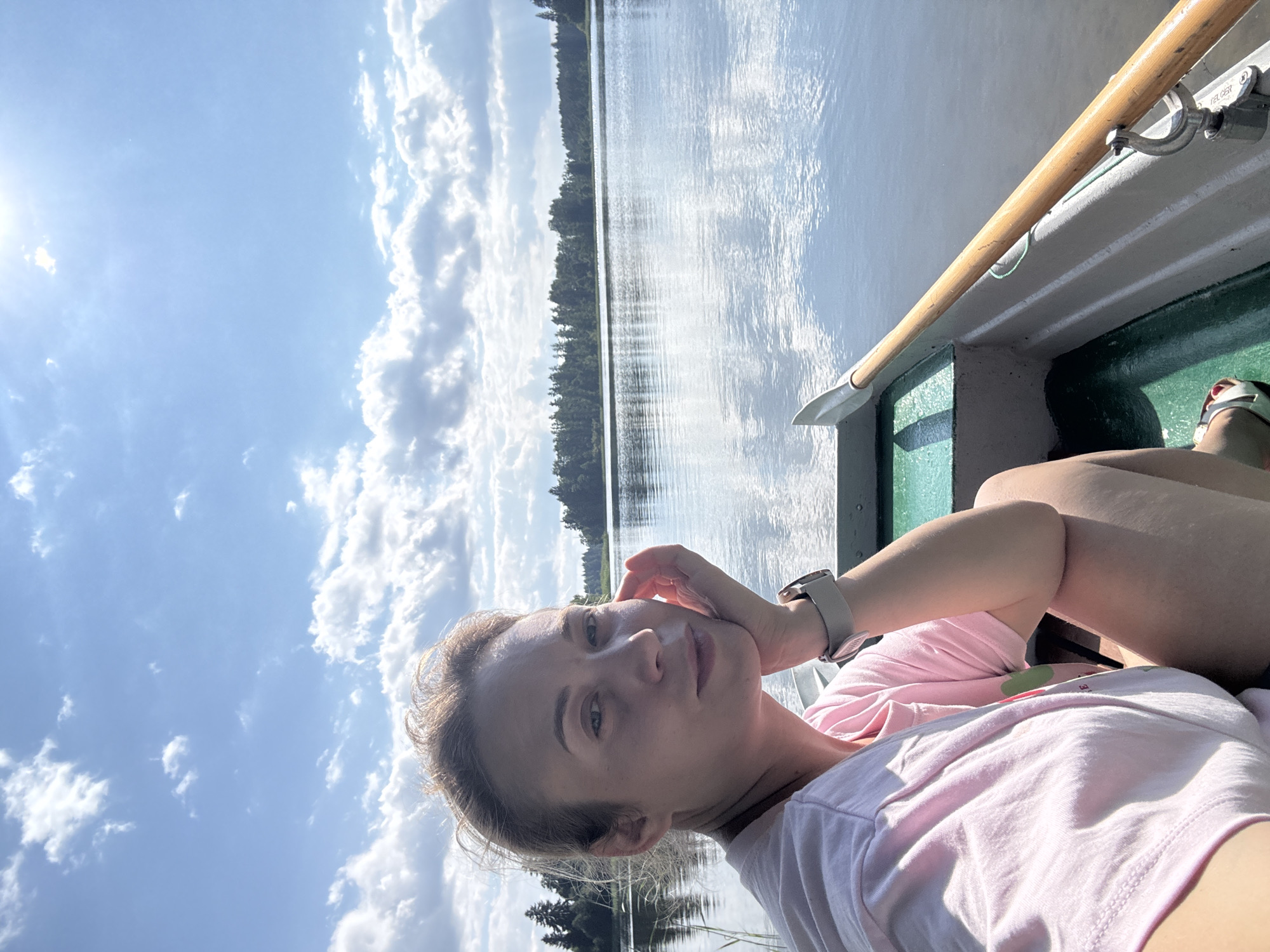 A woman is taking a selfie in a boat on a lake. She has blonde hair and is wearing a pink t-shirt and a watch on her left wrist. The sun is shining and there are white clouds in a blue sky. The water of the lake is calm and reflects the sky and the surrounding trees. There is a wooden oar in the boat with her. The scene appears to be set in the afternoon on a sunny day. The location is Lendži, Latvia.
