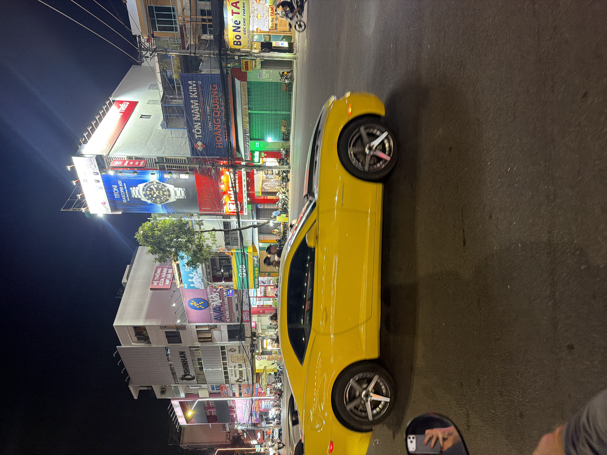 A yellow Chevrolet Camaro is captured in motion on an asphalt street at night, dominating the mid-ground. The vehicle, likely a 5th generation model, features black accents on its hood and a rear spoiler, with its wheels displaying motion blur. In the immediate foreground, a reflection in a side mirror reveals a hand with red nail polish holding a smartphone (identified as an iPhone due to the dual camera bump), indicating the photograph was taken from another vehicle.
The street is flanked by multi-story commercial buildings, illuminated by various signs and streetlights. On the left side of the street, businesses include "SHINHAN," "AVA Kids" (HỆ THỐNG CỬA HÀNG MẸ VÀ BÉ - Mother and Baby Store System), and other establishments with Vietnamese text signage. Numerous motorcycles are parked or moving along the left sidewalk and street.
On the right side of the street, a large billboard for "TITONI SEA SCOPER 600" is displayed on a building. Below it, a blue sign reads "TÔN NAM KIM" (Nam Kim Steel) and "CÔNG TY TNHH HOÀNG QUANG." Further down, a storefront with a green rolling shutter is visible, followed by a business with a yellow sign displaying "BO NE TA" (GÓC CHỢ THANH). Power lines are visible crisscrossing above the street.
Multiple individuals are present on the sidewalks and near the storefronts, some walking or standing, and one person is seen riding a motorcycle on the far right. The scene depicts a busy, brightly lit urban commercial area at night, located in Tay Nha Trang Ward, Vietnam, as indicated by the Vietnamese language on the business signs.