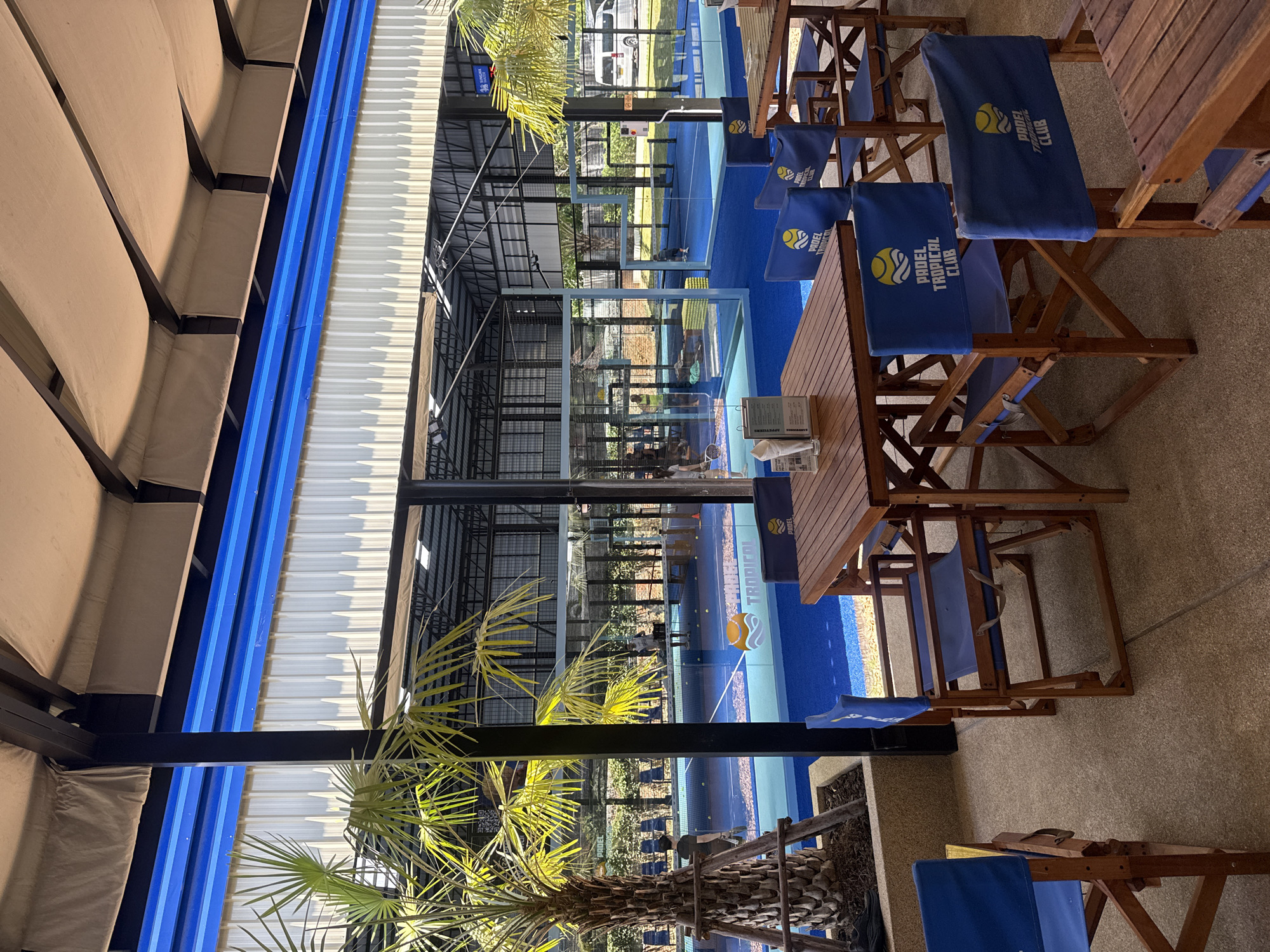 An outdoor seating area overlooks a paddle tennis court with blue turf at the Padel Tropical Club in Ban Bang Rak, Thailand. Several wooden tables and chairs with blue fabric are arranged in the foreground. The paddle court is visible through glass enclosures, with players engaging in a game. Palm trees and lush green vegetation surround the court. In the background, a white van is parked. The sky is clear and sunny.