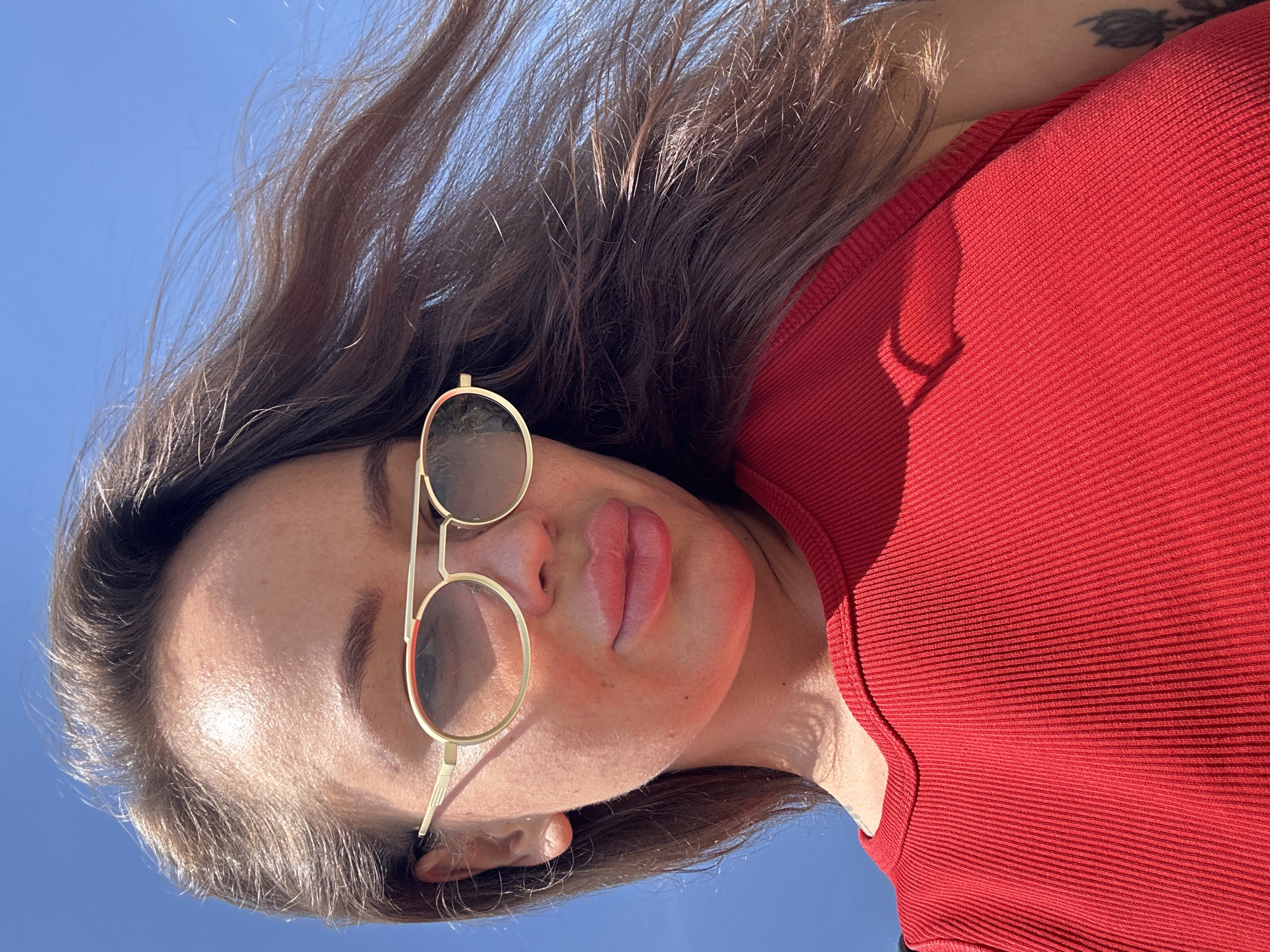 A woman wearing a red top and sunglasses poses for a selfie against a blue sky. Her hair is long and brown, blowing in the wind. The woman's lips are notably full and appear to have been augmented. The image was taken outdoors, likely on a sunny day, given the bright lighting and clear sky. No specific landmarks in Yuzhnouralsk, Russia, are visible.